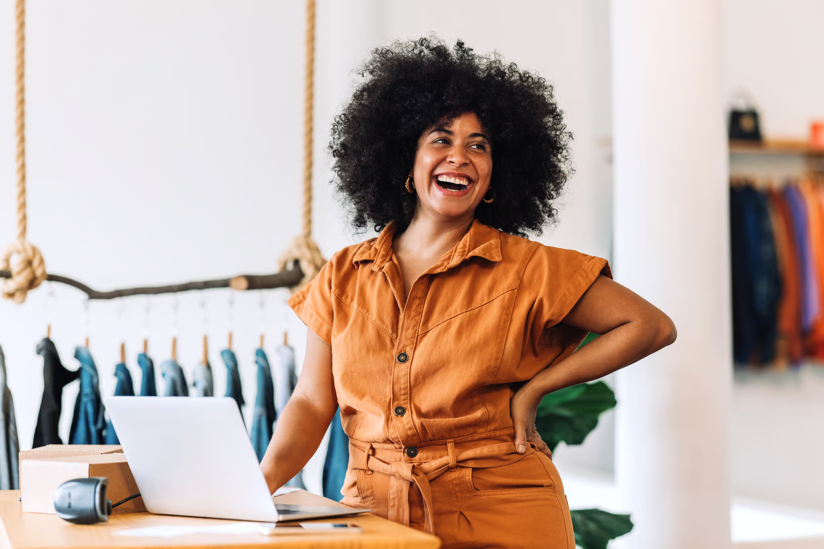 small business clothing shop owner smiling in front of laptop with denim jackets behind