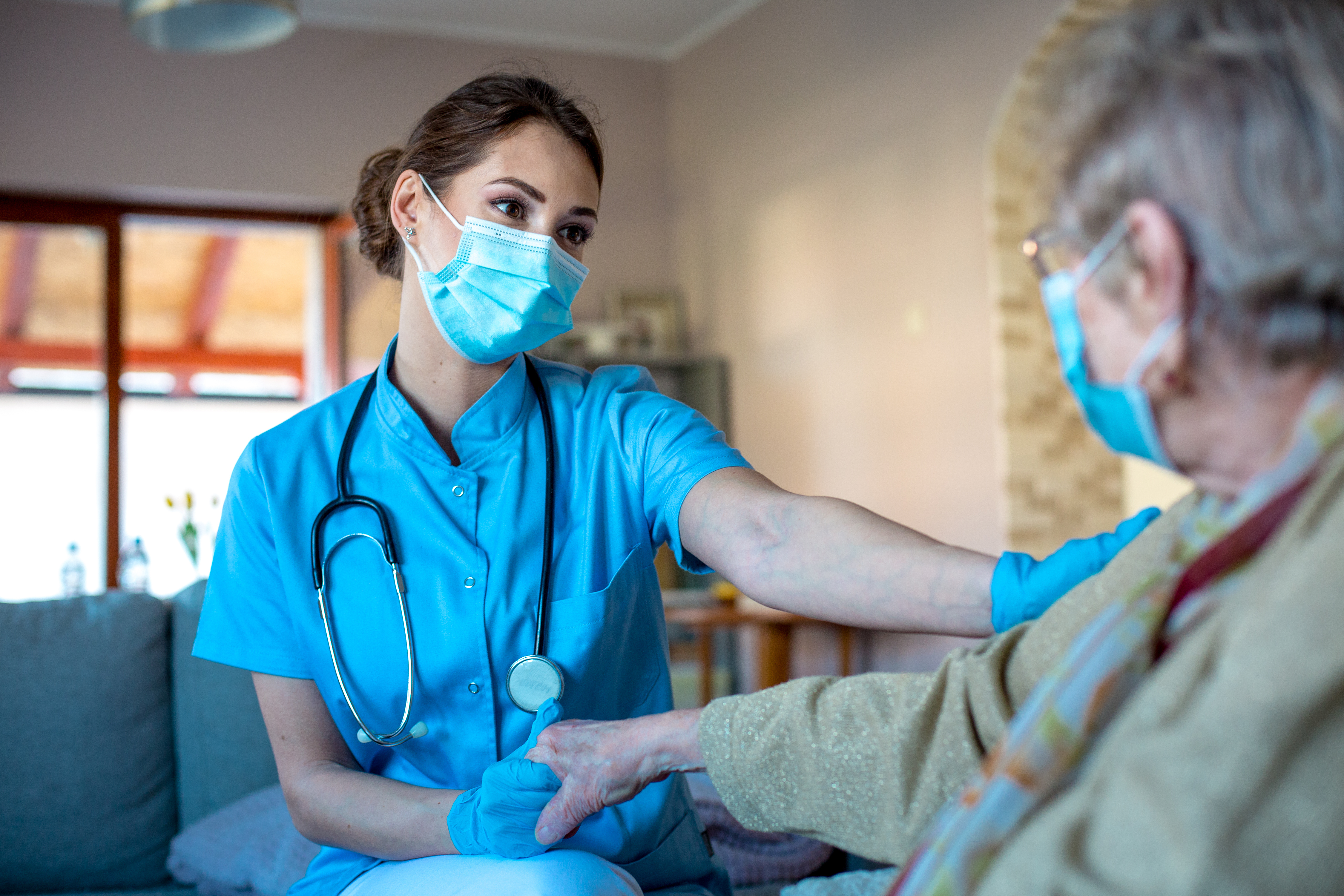 Young nurse is comforting and taking care of her patient. She is holding her hand giving her trust and care that she needs. They are sitting in living room, wearing protective masks.