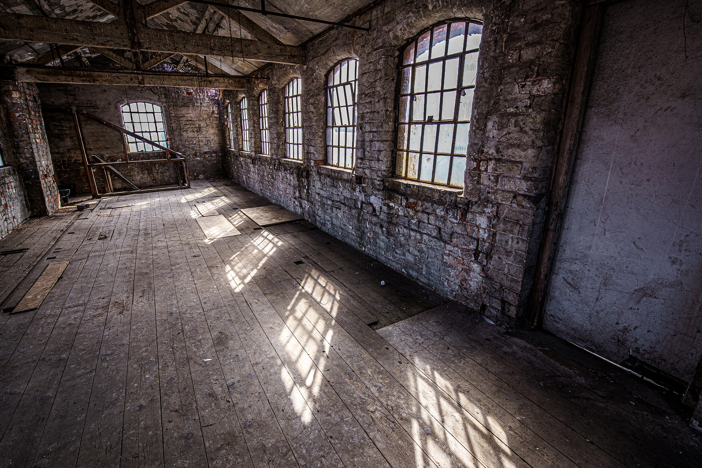 Image of a room in a state of disrepair. There are wooden floorboards and beams, as well as arched windows lining the brick walls. 