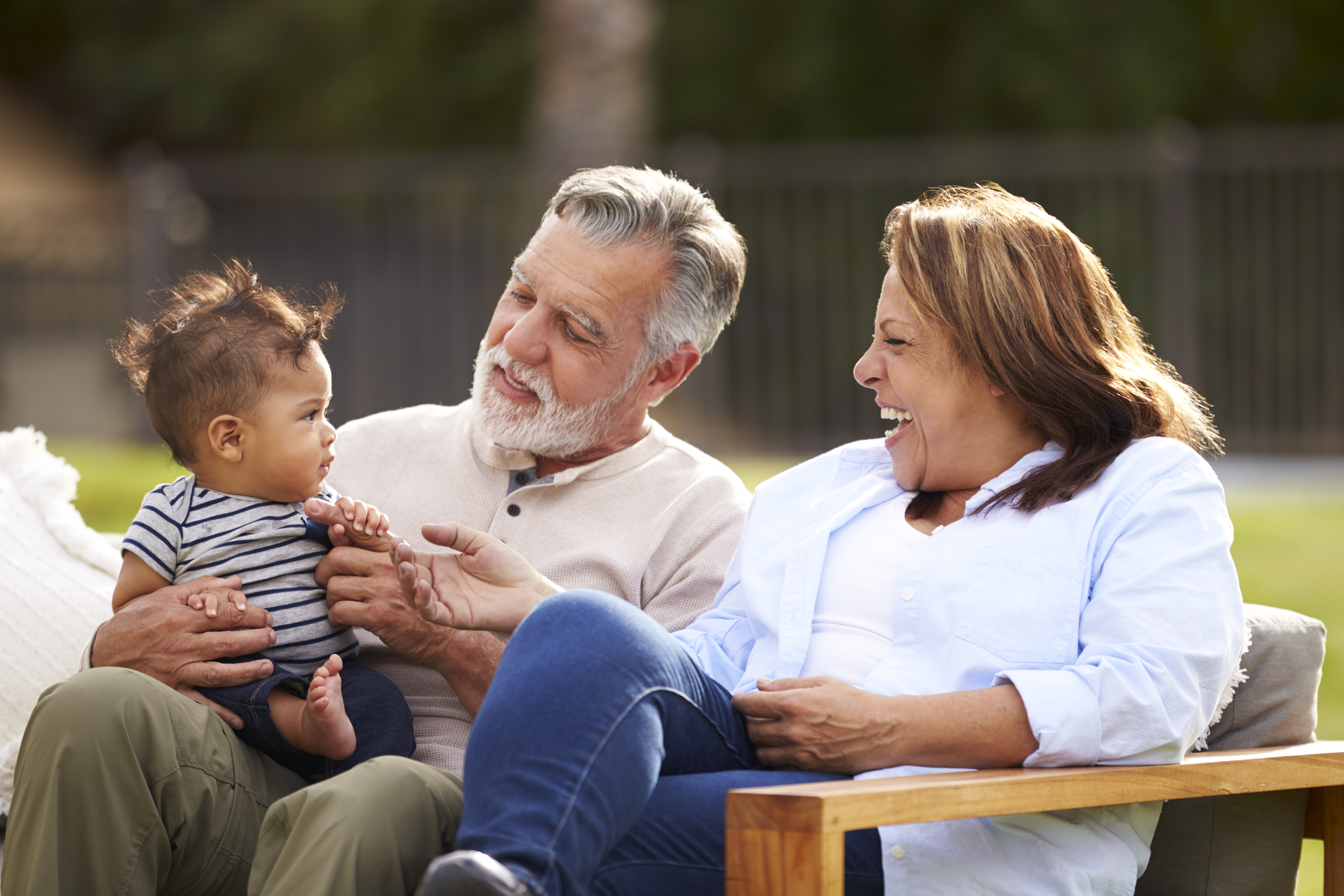 Senior couple sitting in the garden with their baby grandson, smiling at him, front view