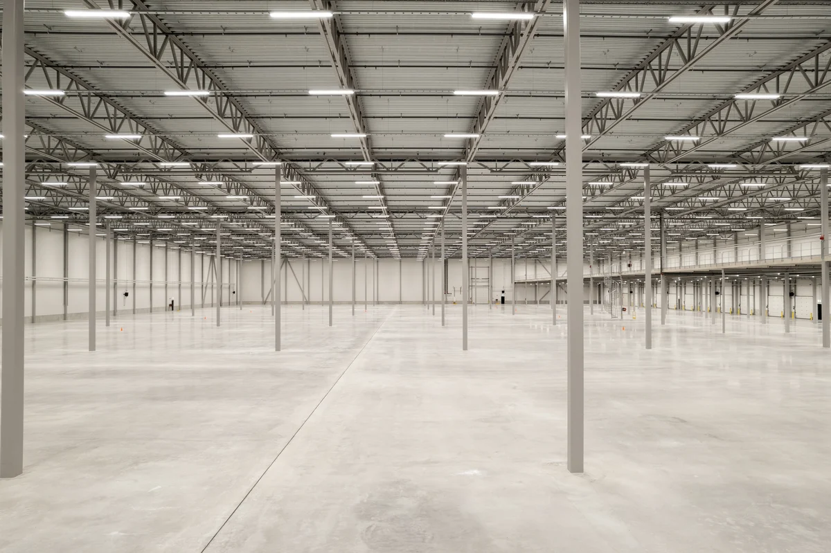 Photograph of a large, empty warehouse interior featuring polished concrete floors, evenly spaced metal support columns, and a high ceiling with exposed beams and fluorescent lighting. The space is well-lit and appears clean and ready for storage or industrial use, with no equipment or materials present.