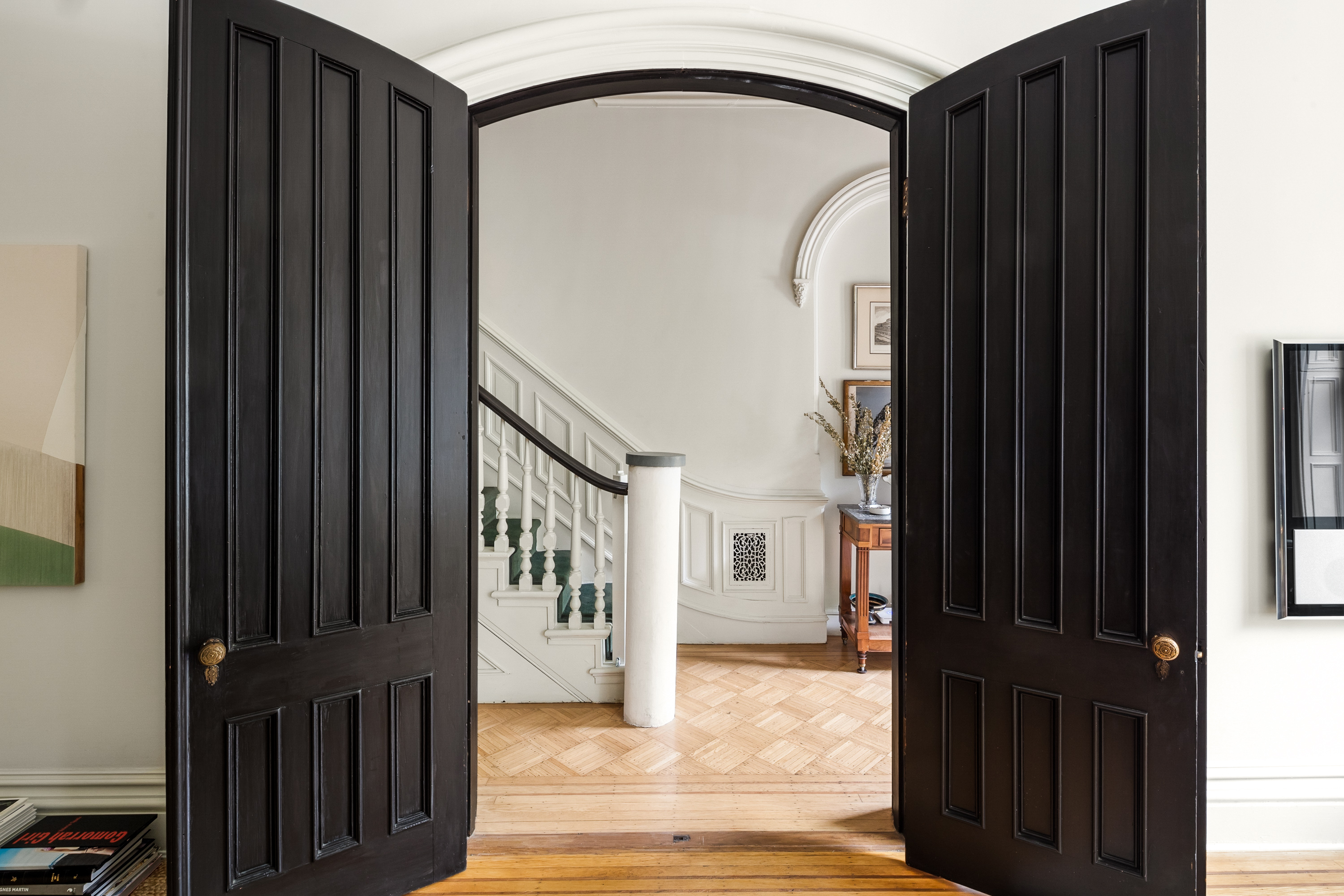 A pair of open black wooden doors reveal a hallway with a staircase that has white railings and green balusters, leading to an upper floor; the space features light-colored walls, wooden flooring, and a decorative arch above the doorway.