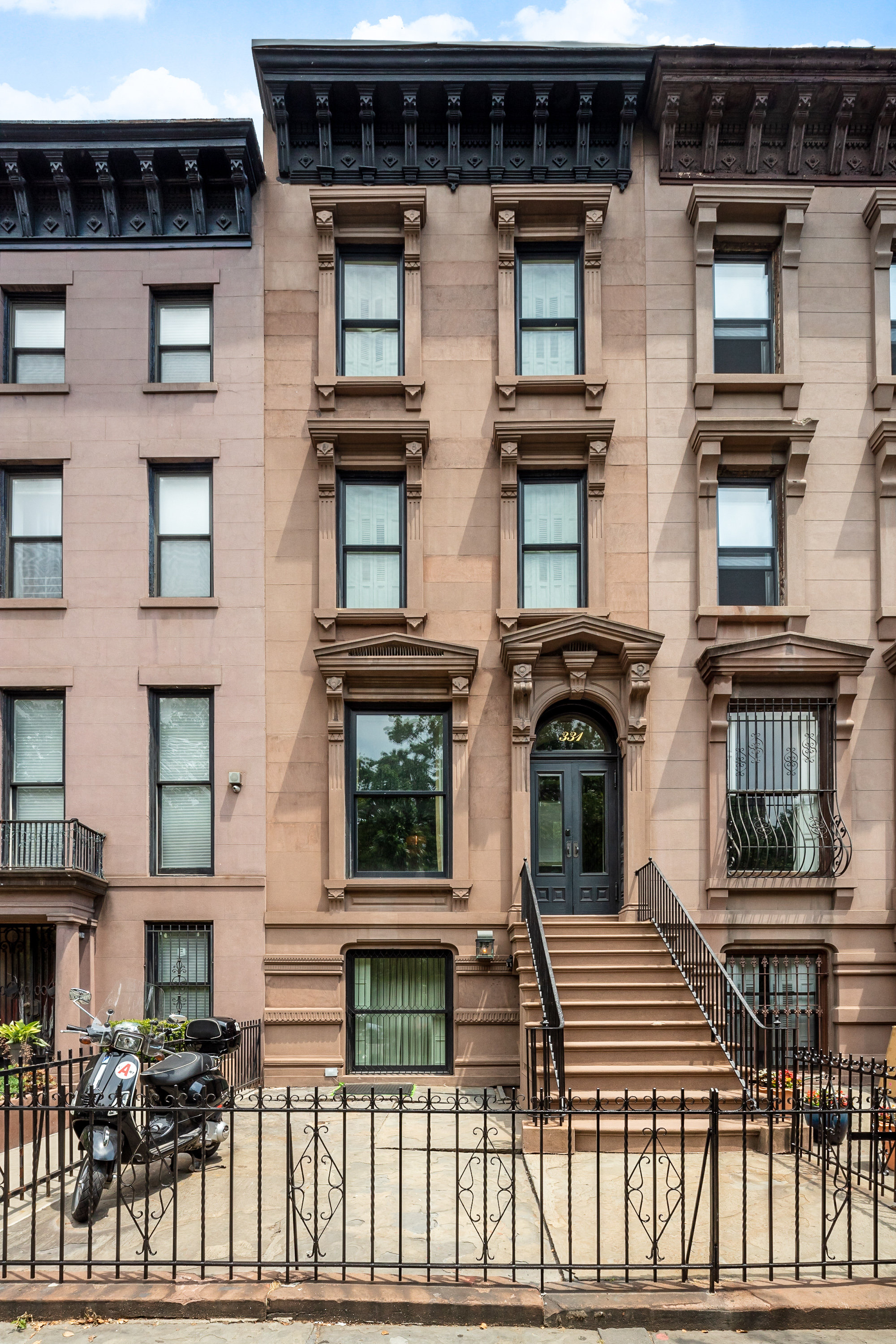 A three-story brownstone townhouse with large windows, decorative moldings, a black iron fence, and a staircase leading to the front door; a motorcycle is parked to the left of the entrance.