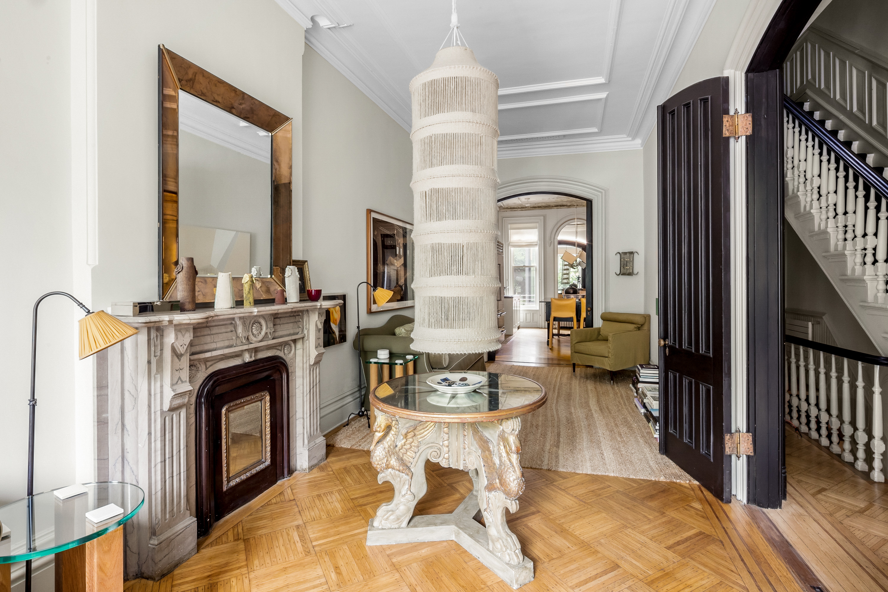 A spacious living room with a large chandelier, a decorative fireplace with a mirror above it, an elegant wooden table in the center, parquet flooring, arched doorways, and a staircase on the right.