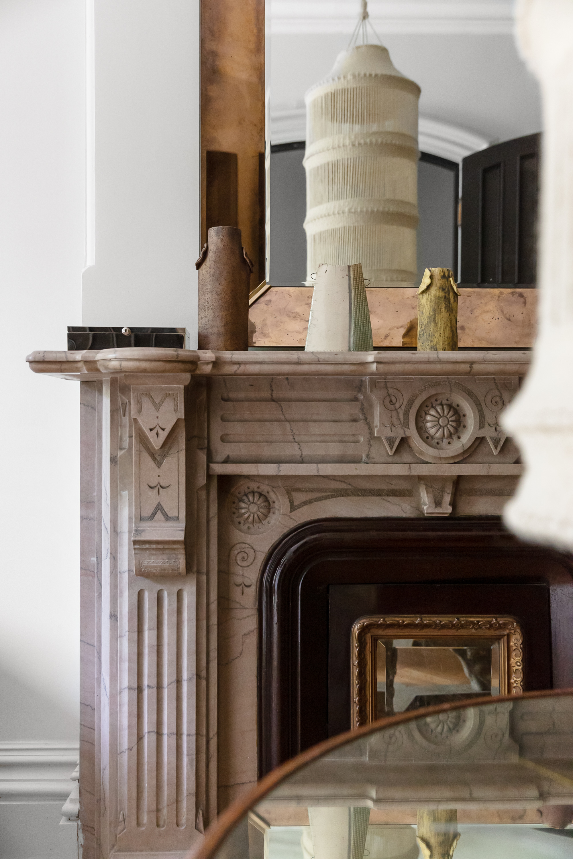  A marble fireplace mantel with detailed carvings, decorated with three assorted vases, a mirror above reflecting a hanging light fixture, and part of a round glass table visible in the foreground.