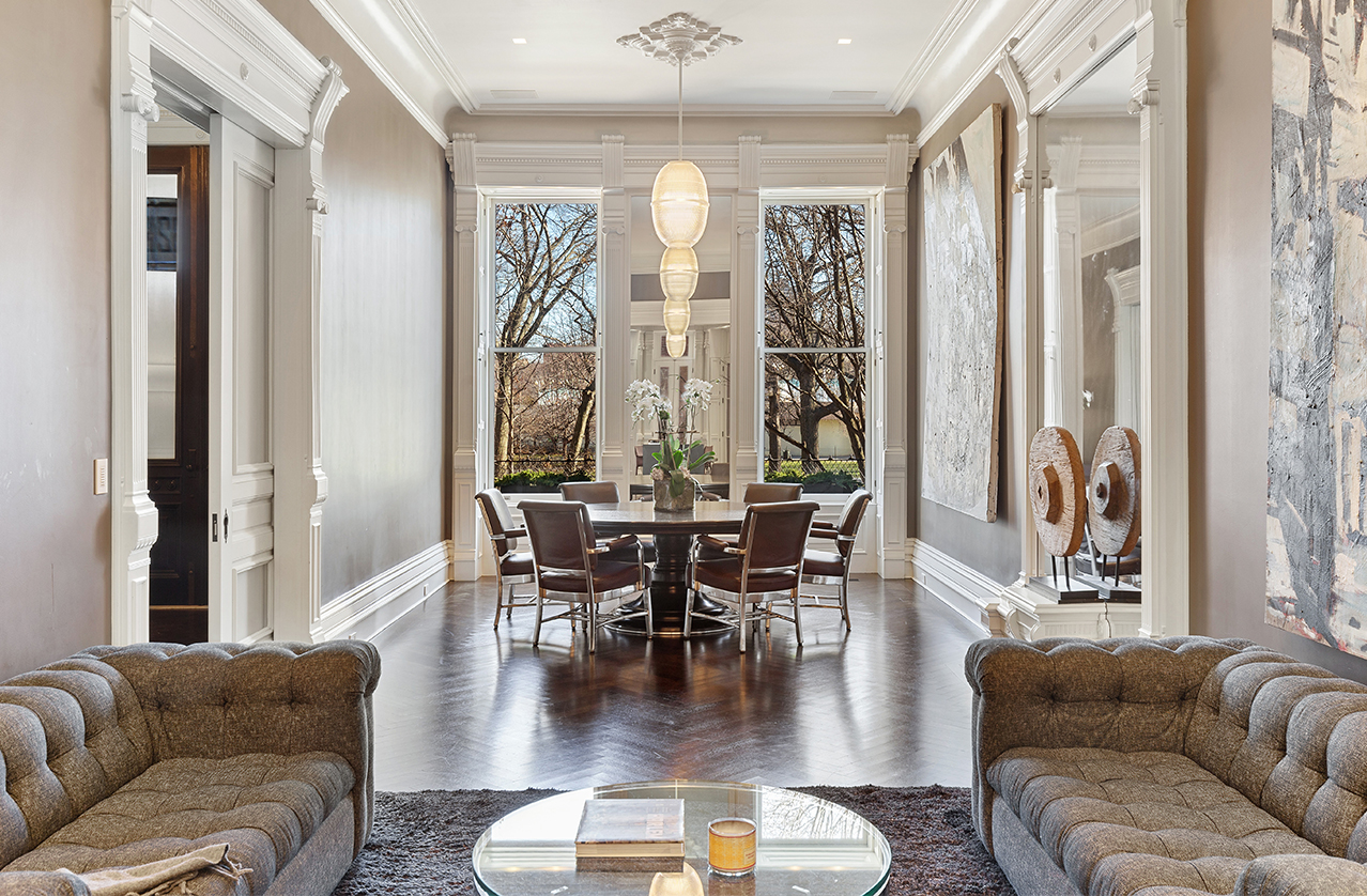  A stylish living room with two gray sofas, a round glass coffee table, and a dining area in the background, featuring large windows, dark wood floors, and modern decor.