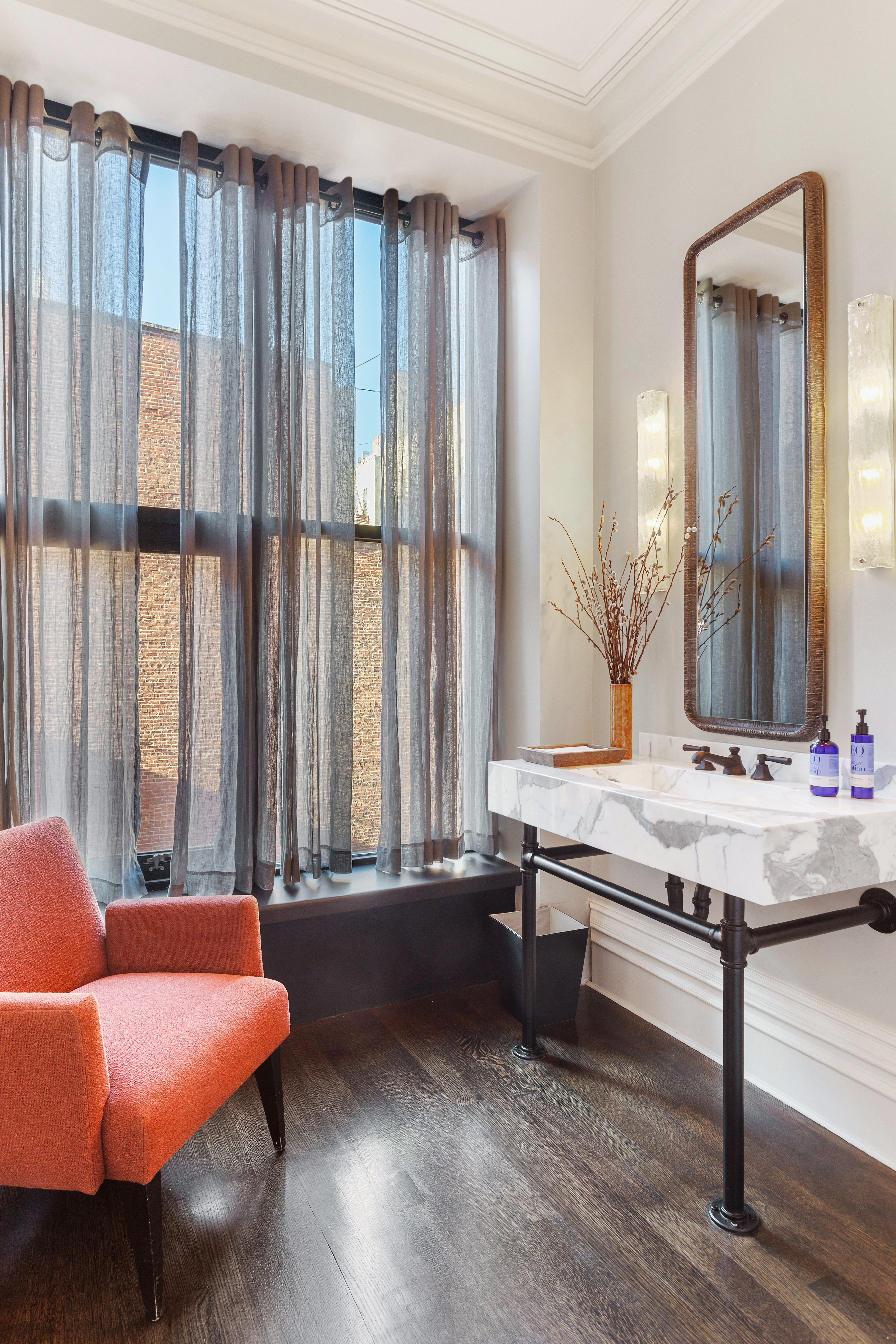 A modern bathroom with a white marble sink, a red-orange armchair by a large curtained window, and dark wood flooring with soft lighting.