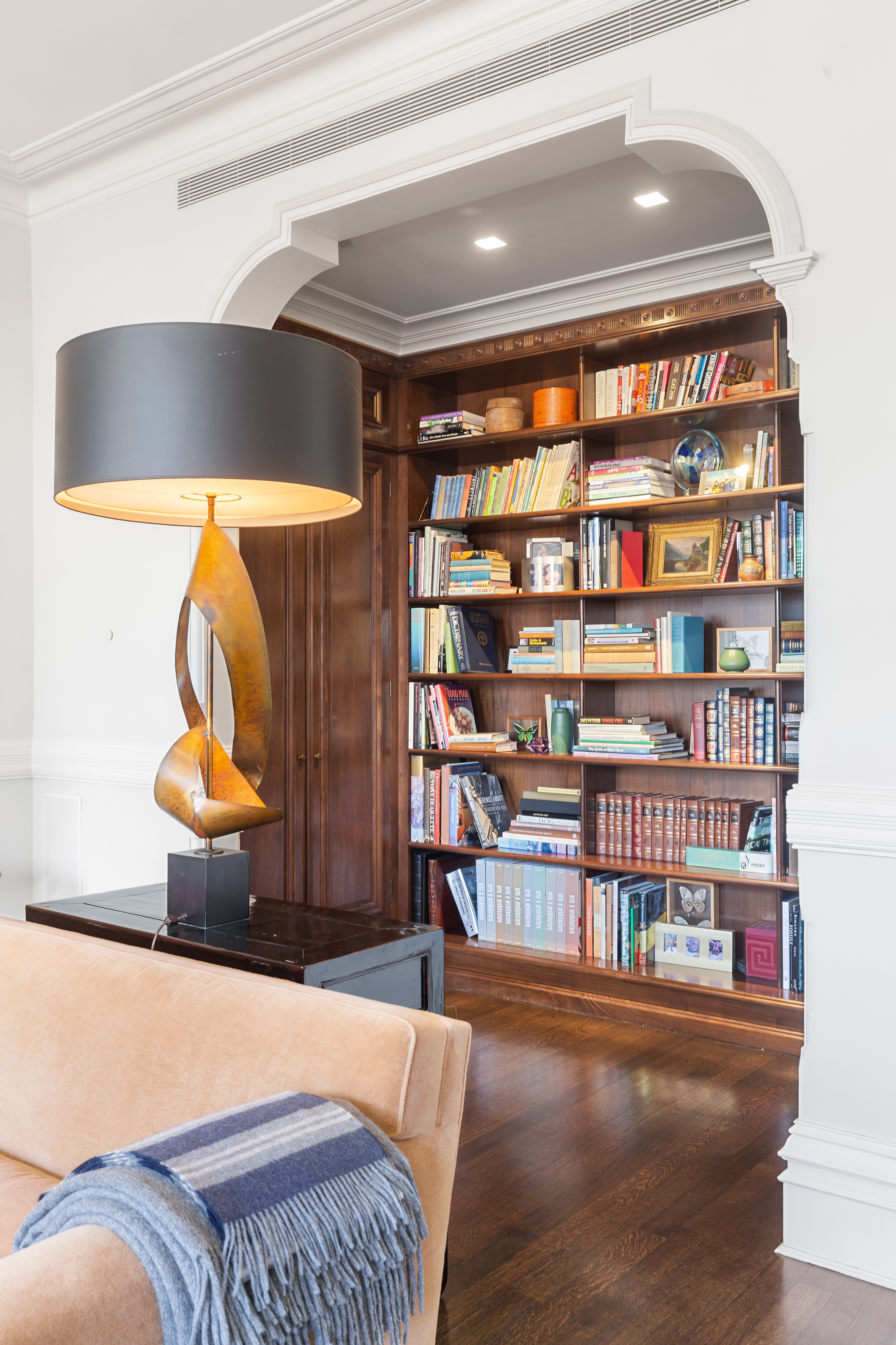 A cozy living room with a beige sofa, a large bookshelf filled with books and decor, a modern lamp, and wooden flooring with white walls and molding.