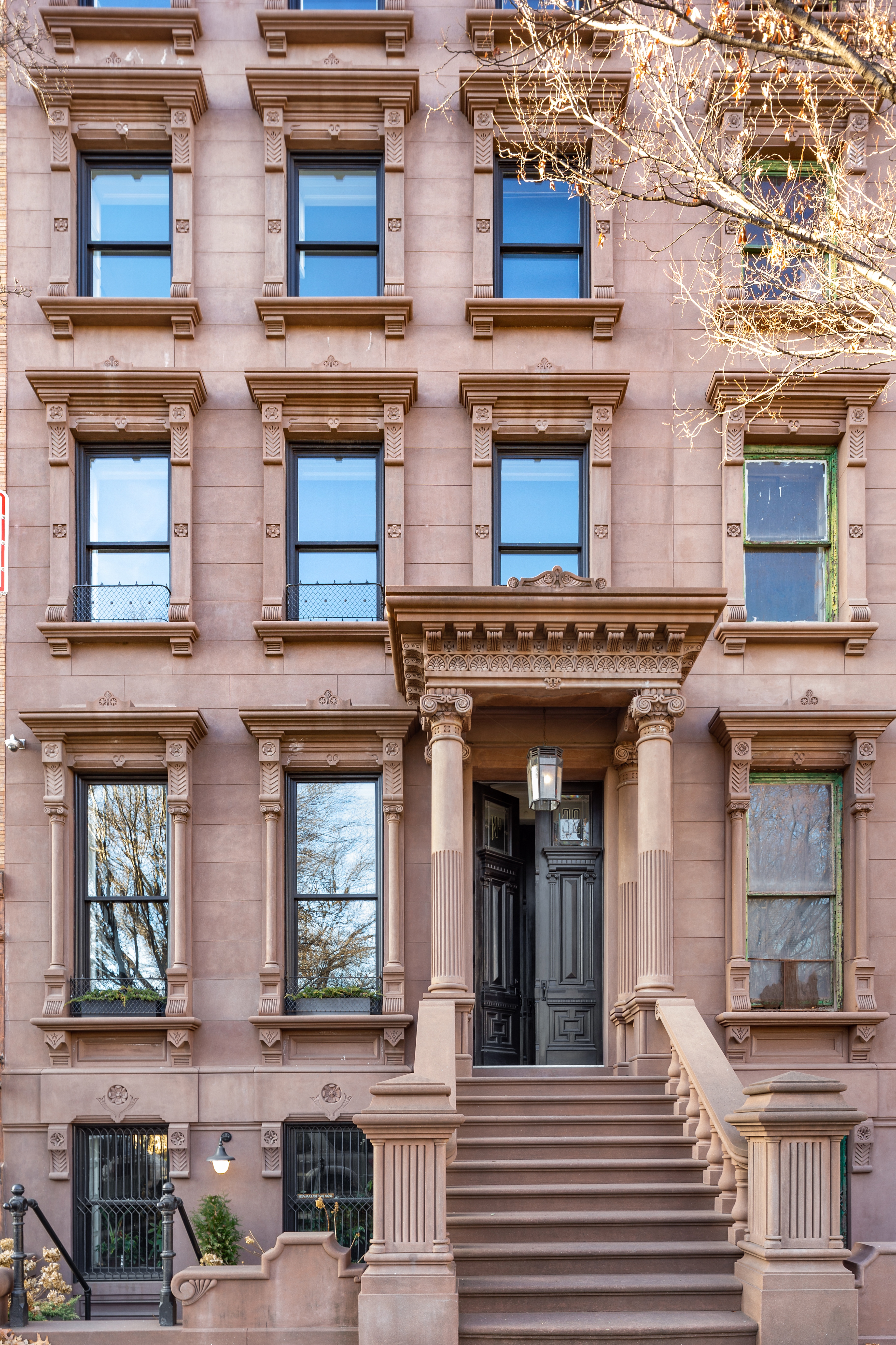 The exterior of a three-story brownstone building with large windows, decorative columns, and a staircase leading to double doors under an ornate portico.