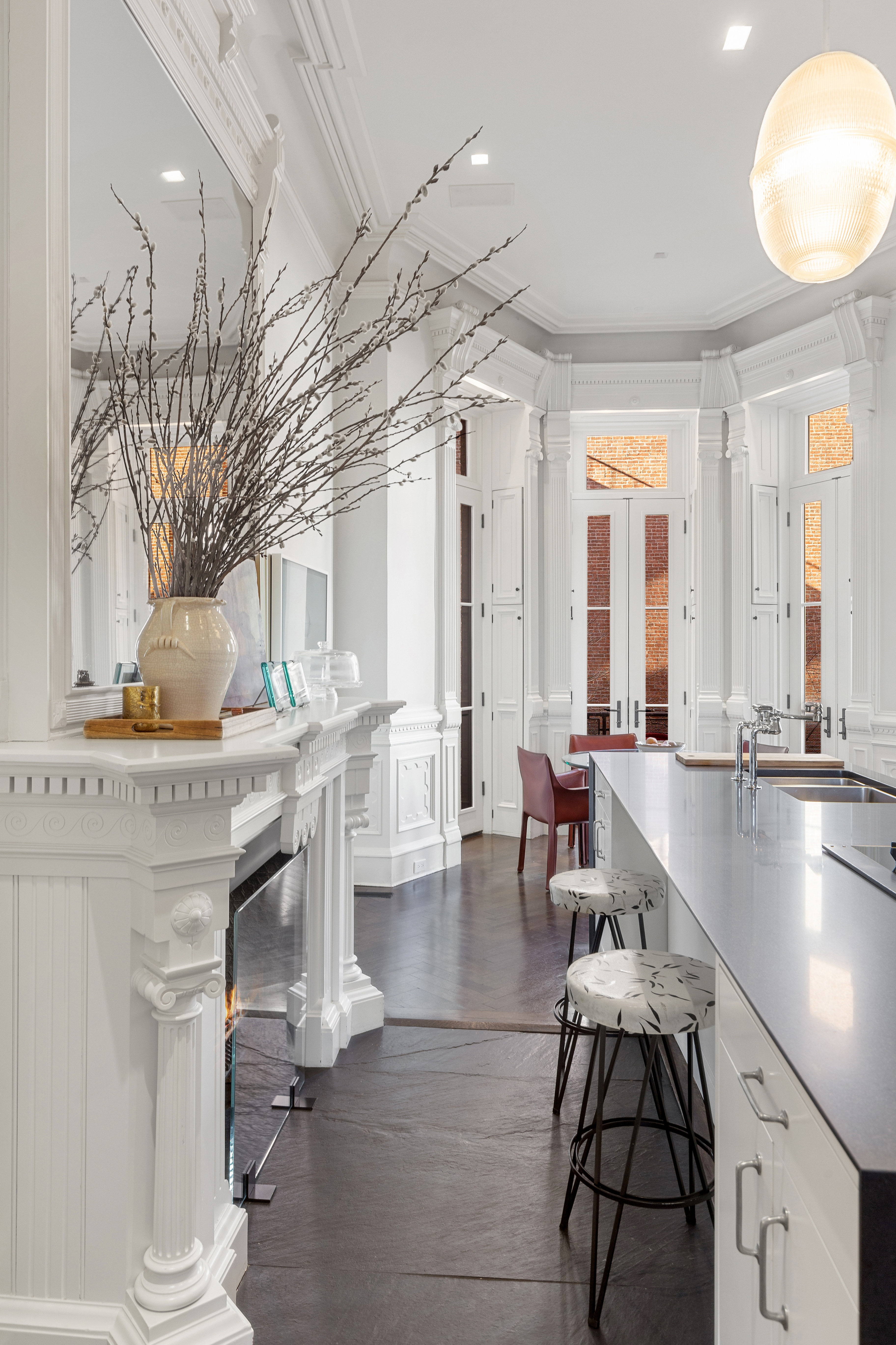 A modern kitchen with white cabinetry, a large island with a dark countertop and bar stools, high ceilings with molding, and a red chair near sunlit windows.