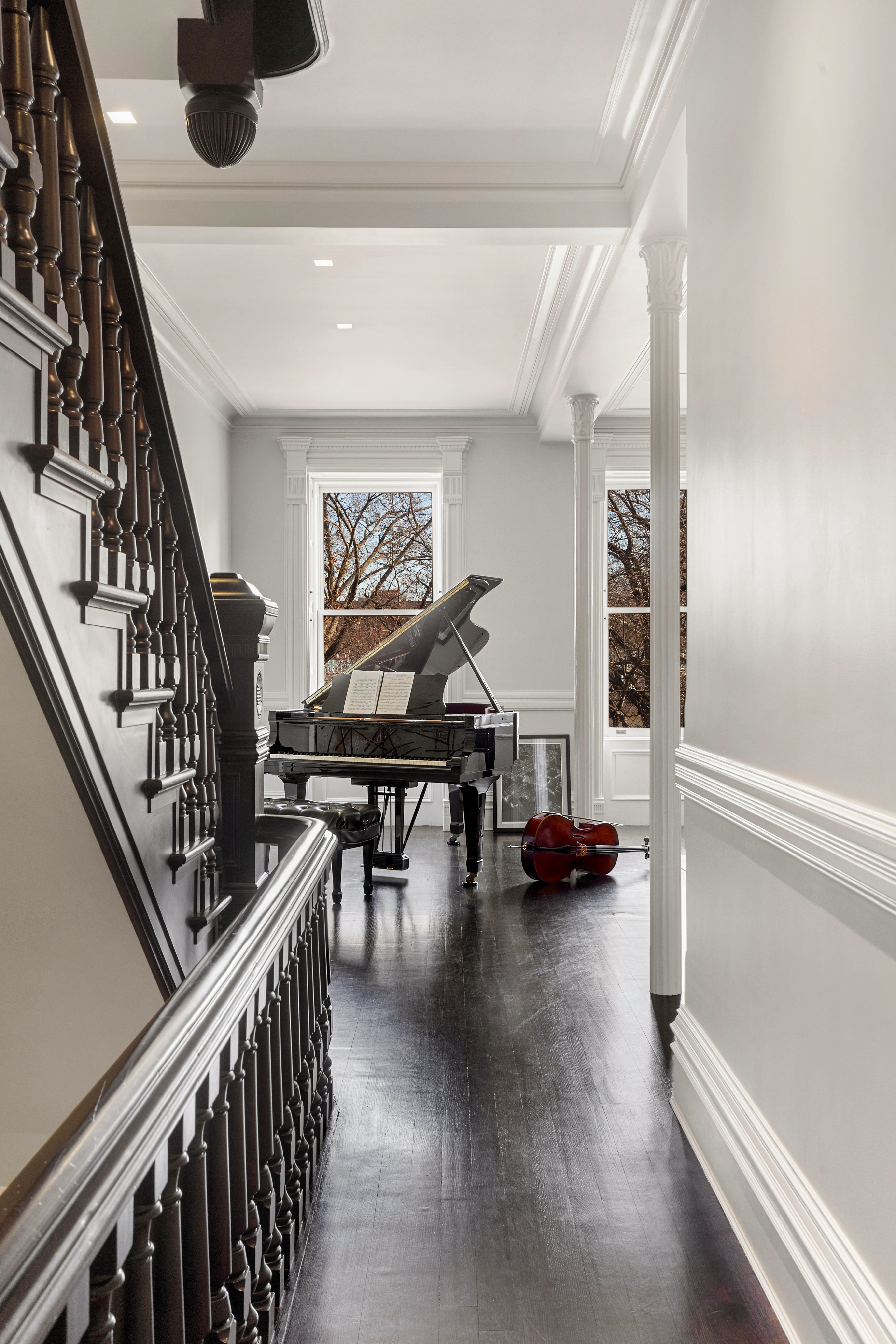 A hallway with a staircase leading to a room featuring a grand piano with sheet music and a cello, set against white walls and dark wood flooring.