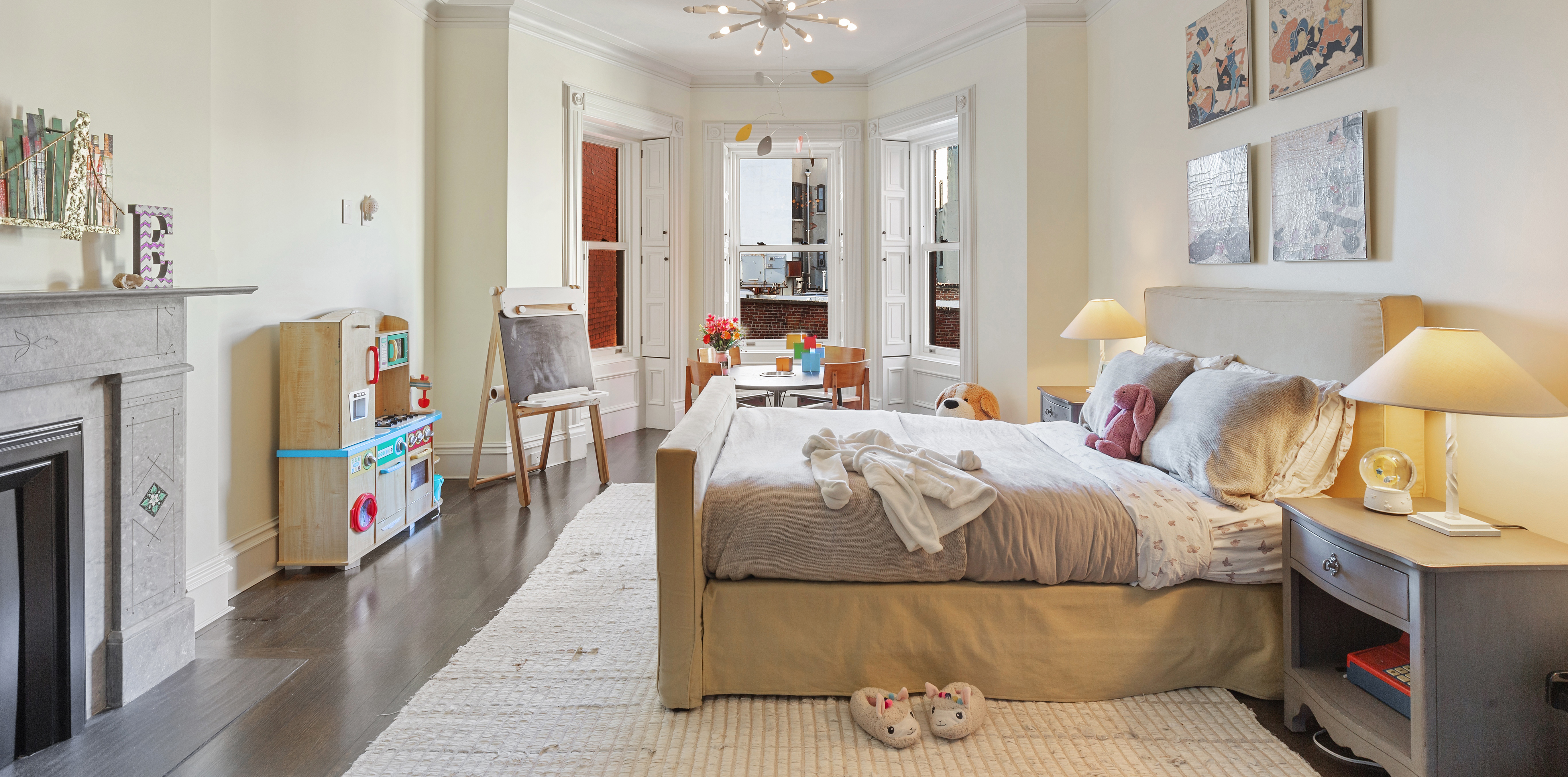A spacious bedroom with a large bed, fireplace, children's play area, and wooden flooring, featuring nightstands with lamps and artwork above the bed.
