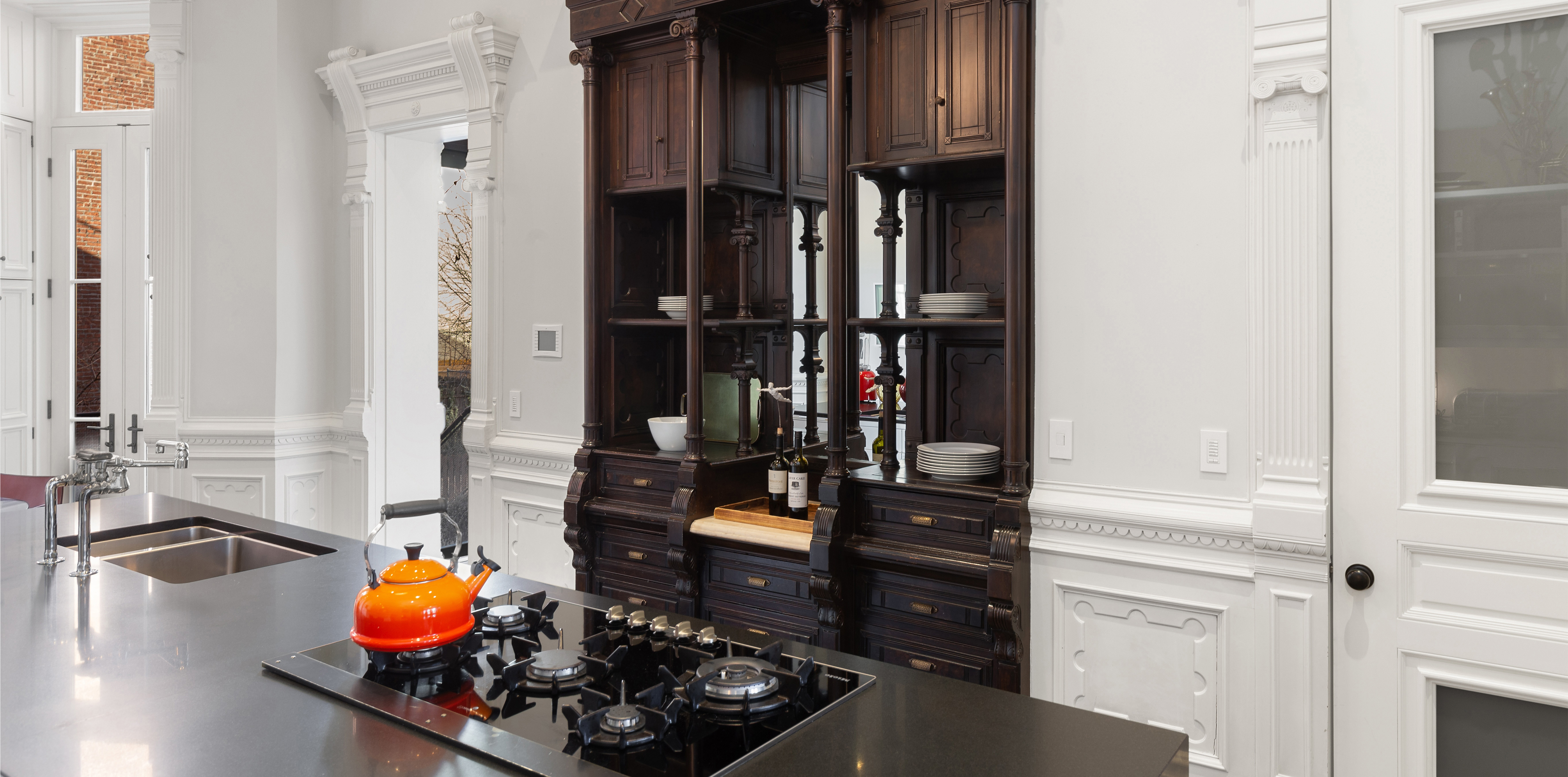 A modern kitchen with a large island featuring a black countertop, built-in gas stove with an orange kettle, and an ornate dark wooden cabinet in the background.