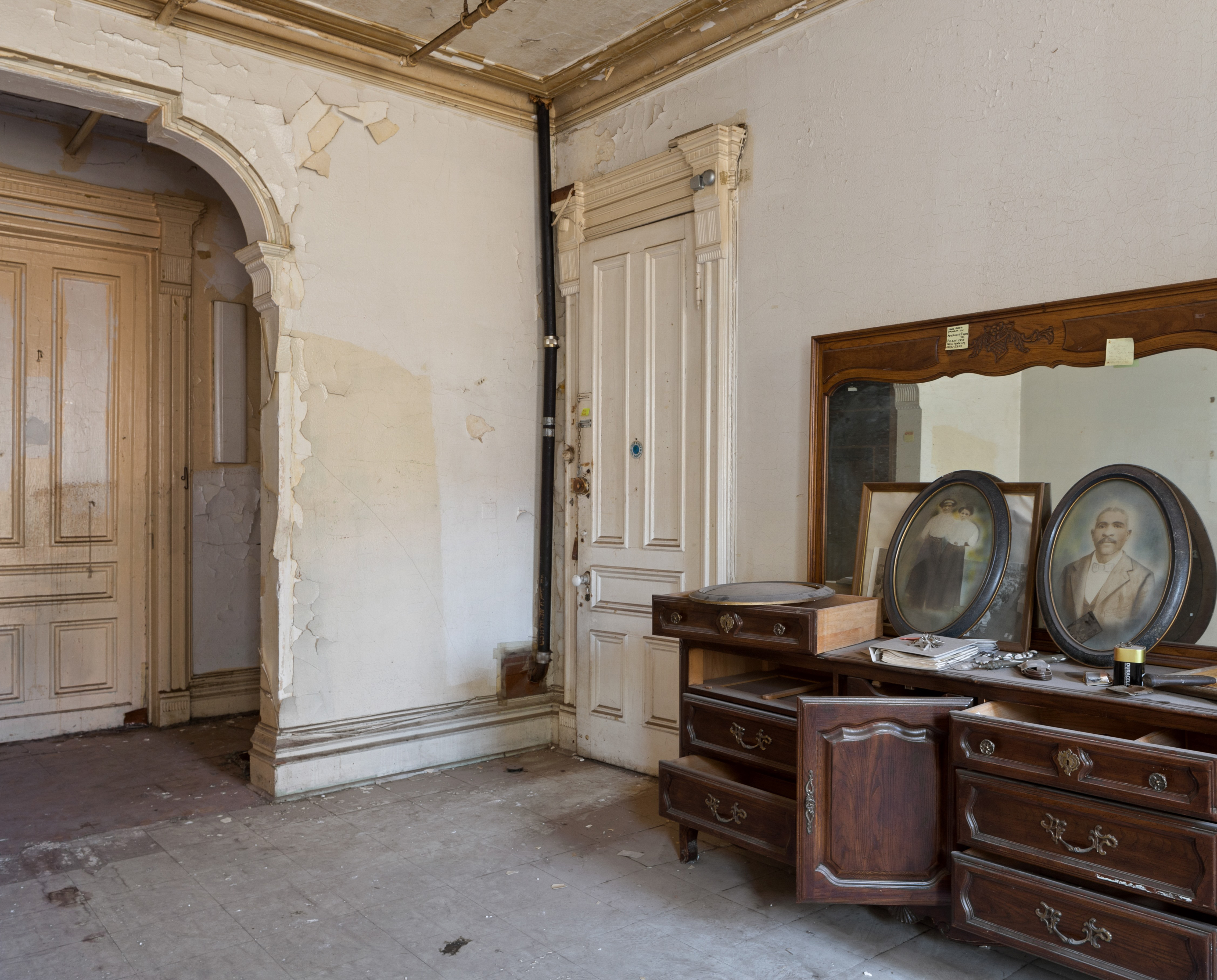 Wooden dresser with a large mirror and two framed portraits, set in a room with an arched doorway and wooden doors.
