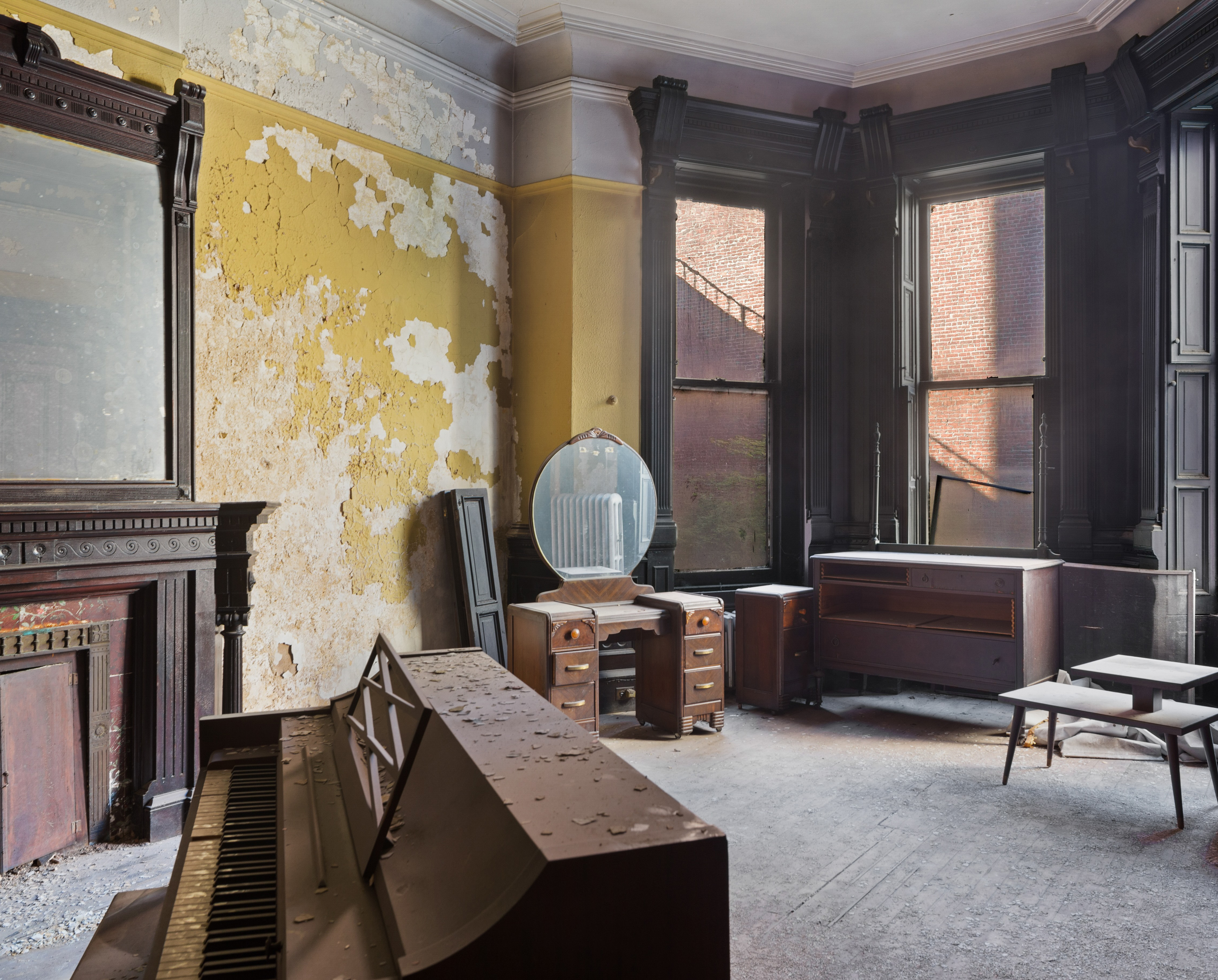 Room with a piano, ornate fireplace topped by a mirror, a desk with drawers and chair, two small tables, and large windows letting in natural light.