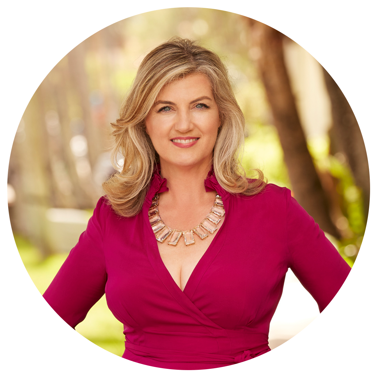 A person with shoulder-length blonde hair wearing a magenta dress and a large statement necklace stands outdoors with a background of blurred trees and greenery.