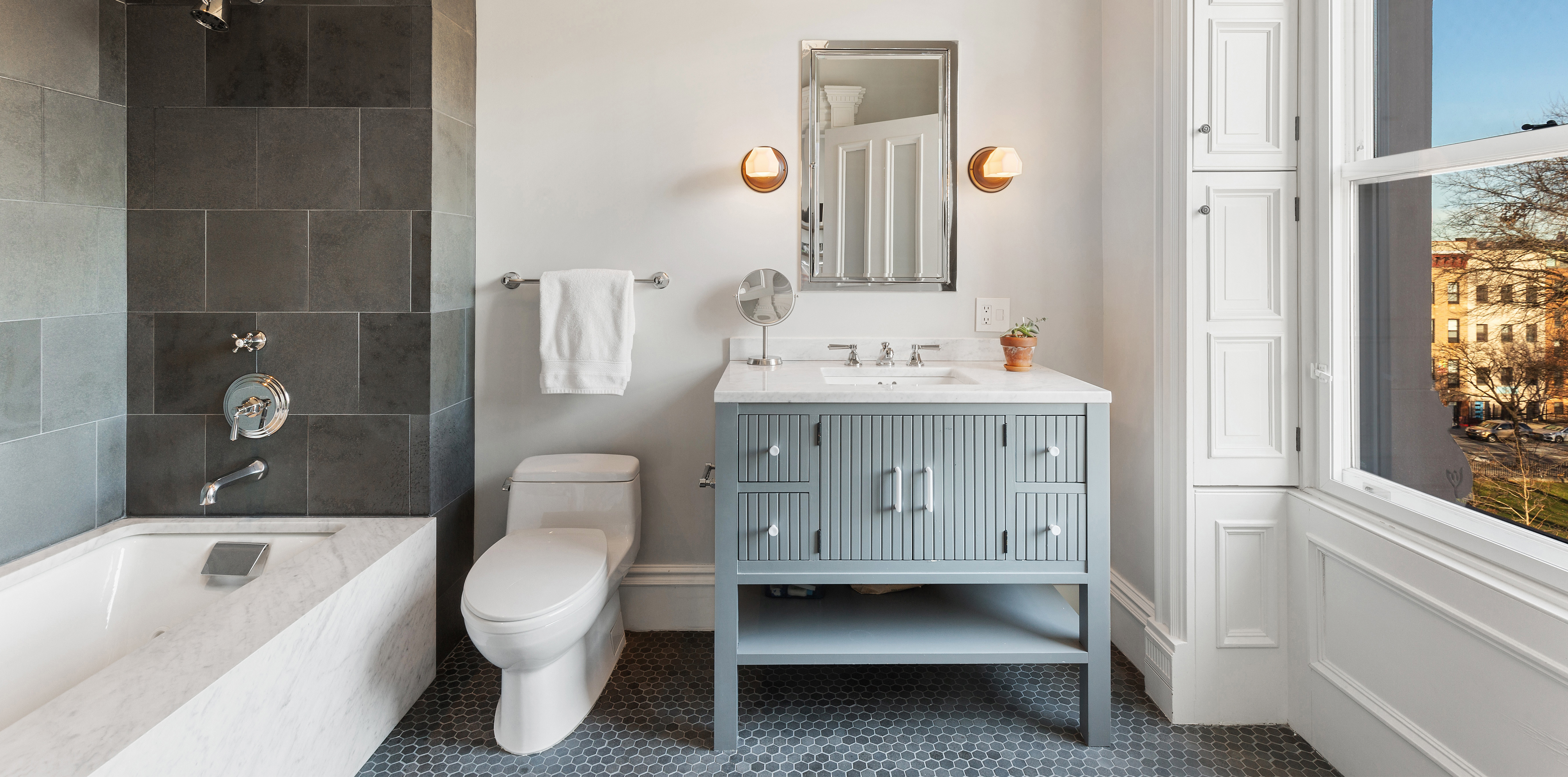 A modern bathroom with a bathtub, light blue vanity, patterned tile floor, and a large window offering a view of buildings and trees.