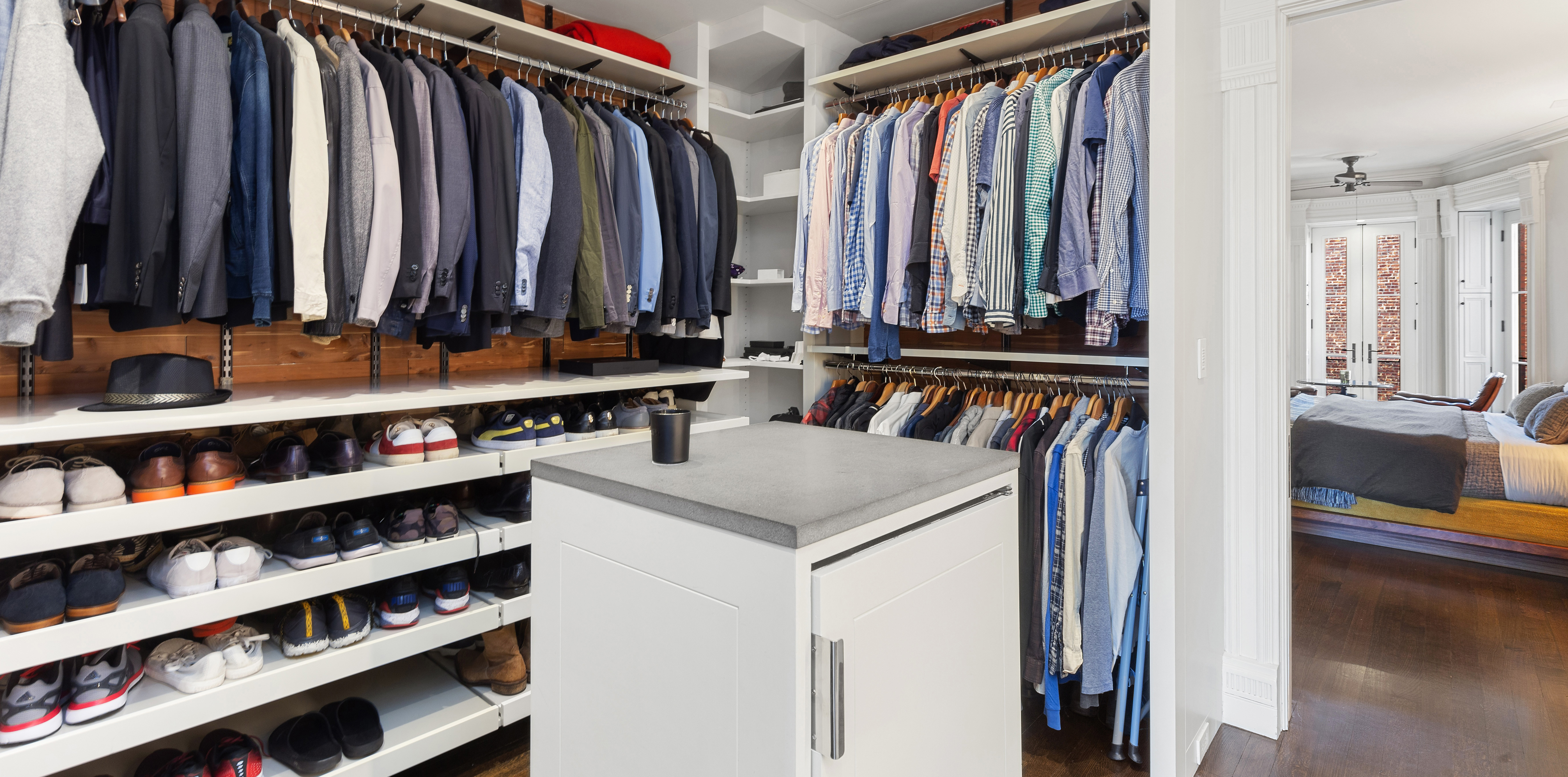 A spacious walk-in closet with neatly arranged clothing, shelves for shoes and hats, and a central island with drawers, opening into an adjacent bedroom.