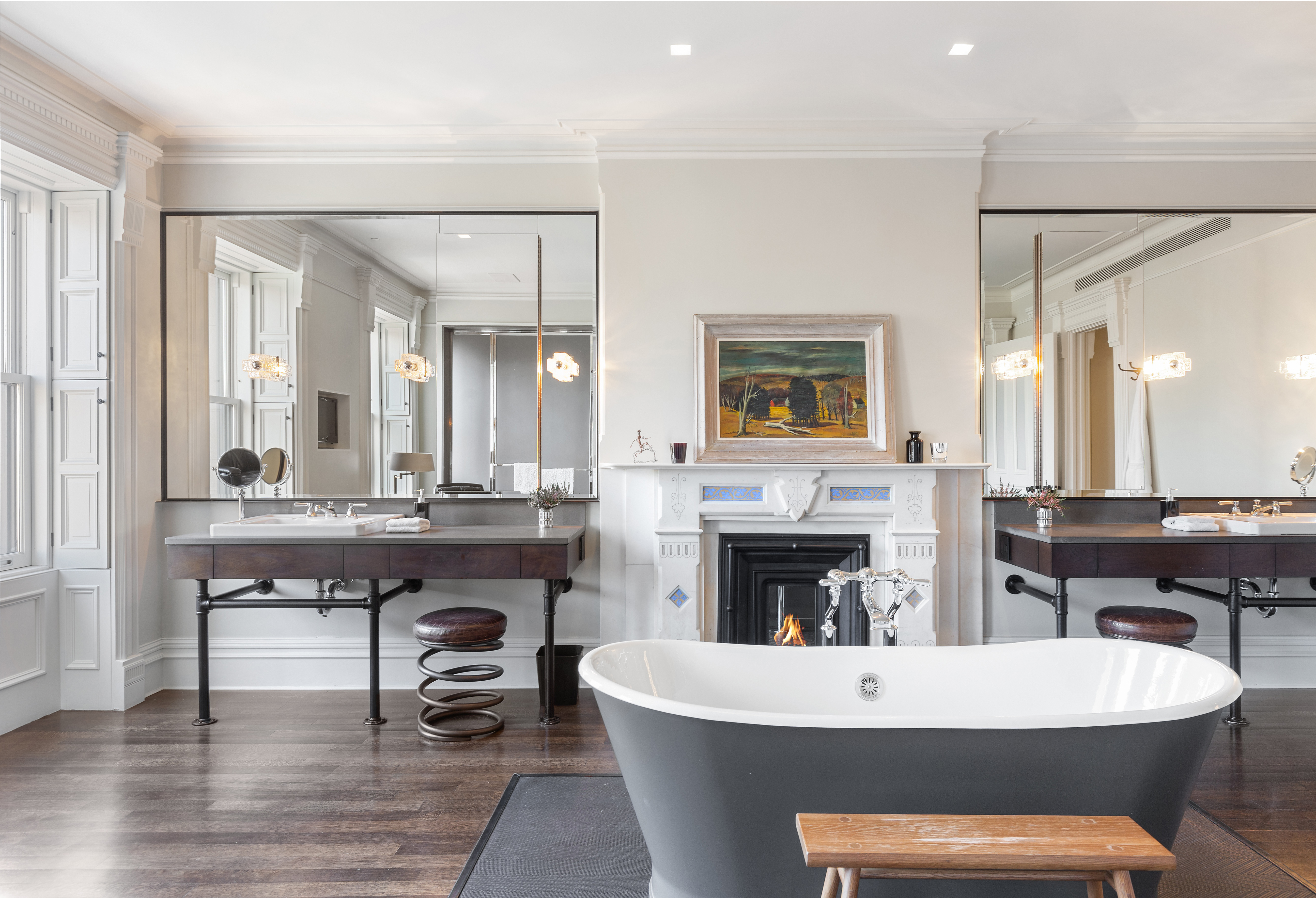 A spacious bathroom with a freestanding cast-iron and ceramic bathtub, two large mirrors above double sinks, a fireplace with artwork above it, and wooden floors.
