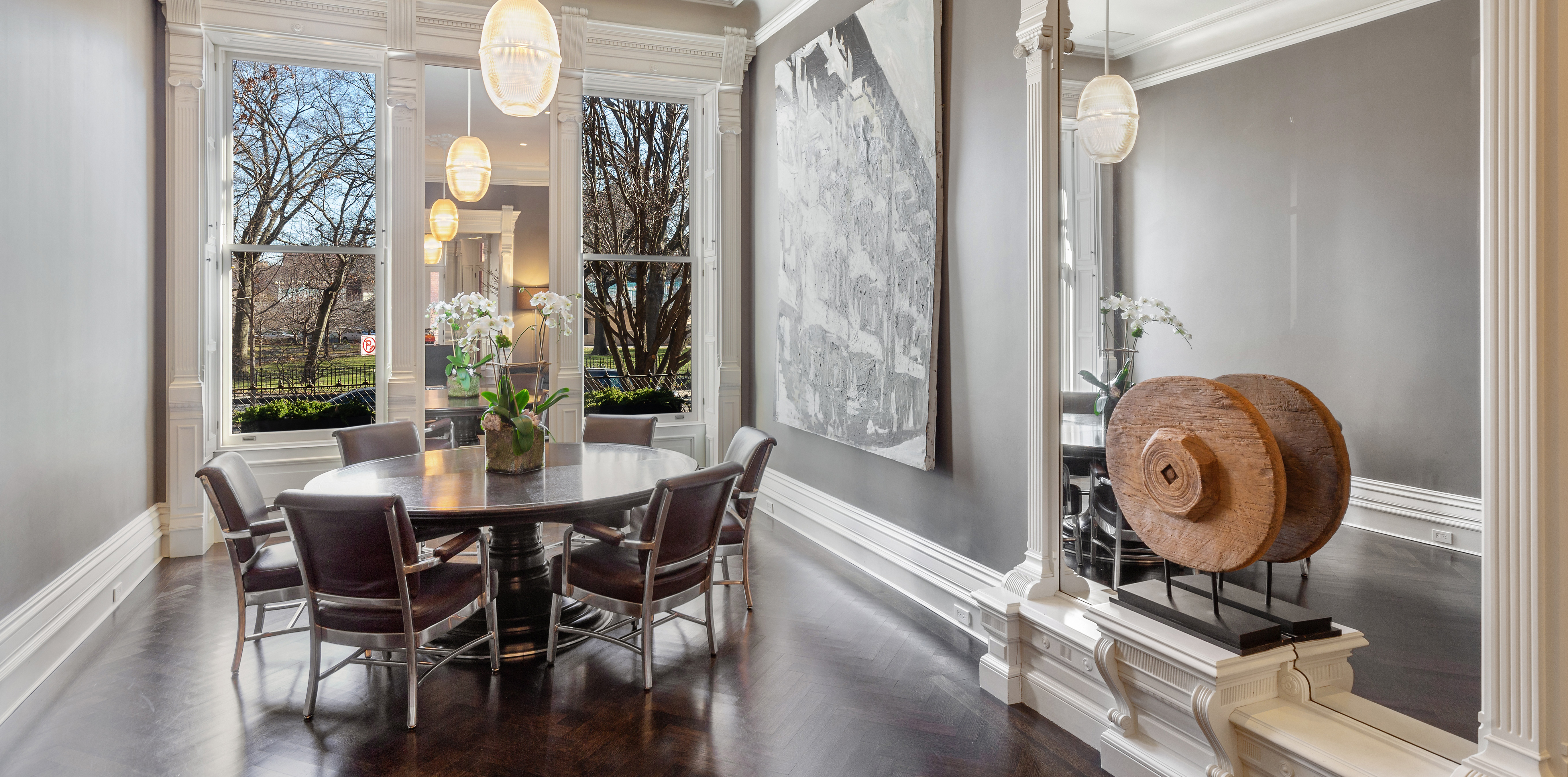A dining room with a round wooden table, six chairs, large windows, pendant lights, and a decorative wooden wheel near a wall with artwork.