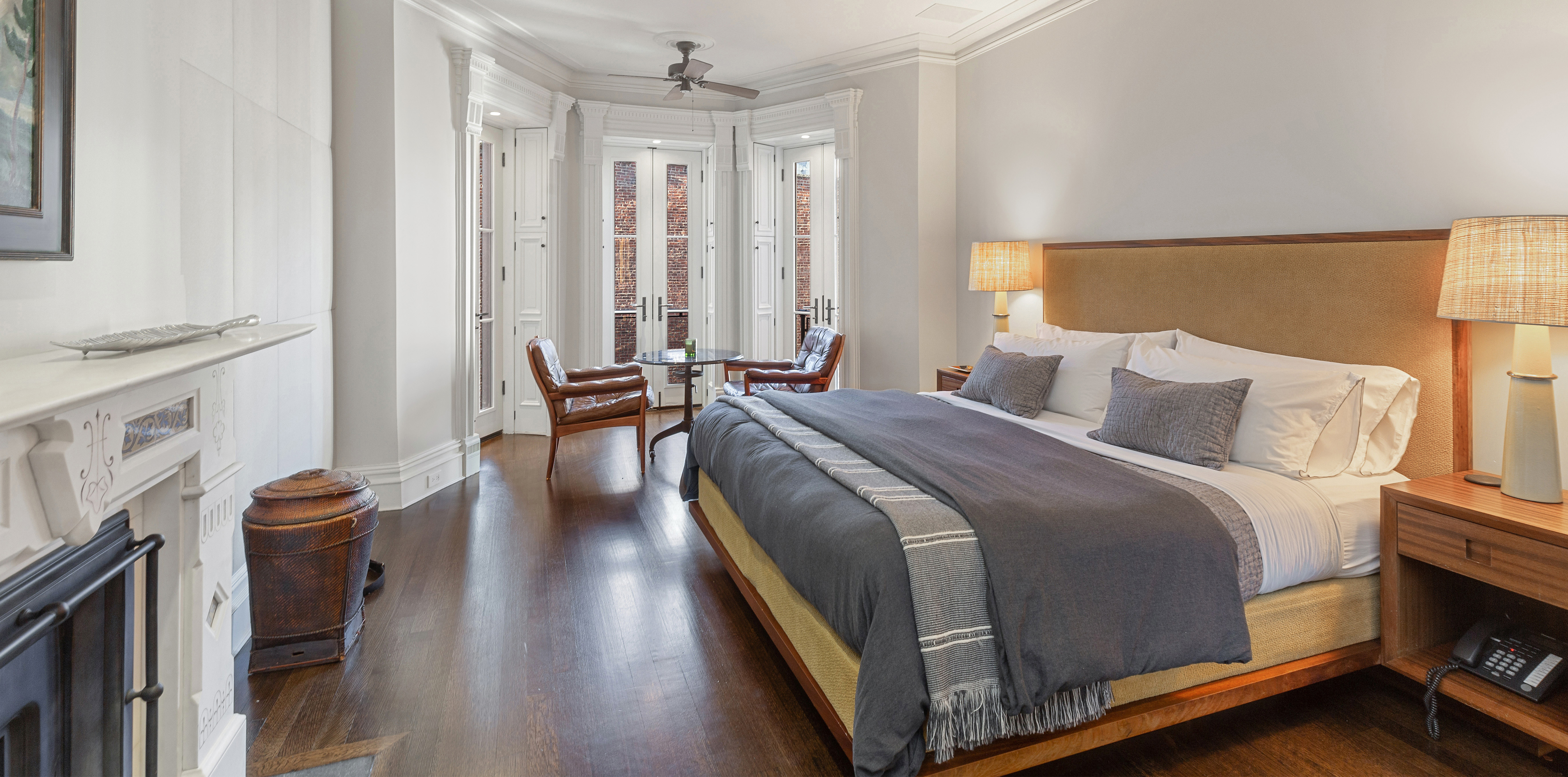 A spacious bedroom with a large wooden-framed bed, dark wood floors, a fireplace, and two chairs by large windows that fill the room with natural light.