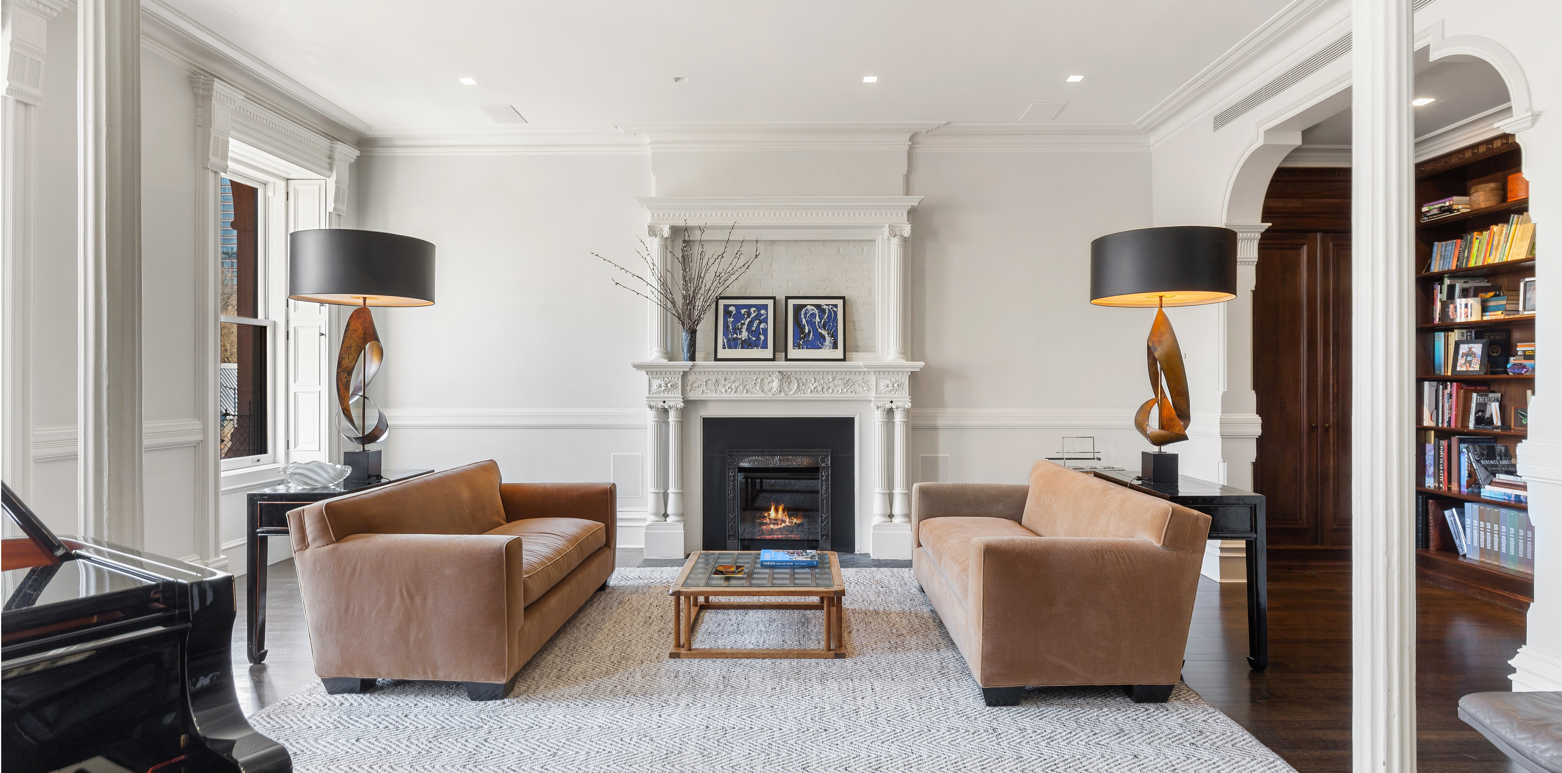 A living room with two brown leather sofas, a lit fireplace, framed artwork, a coffee table, and a grand piano near a window.