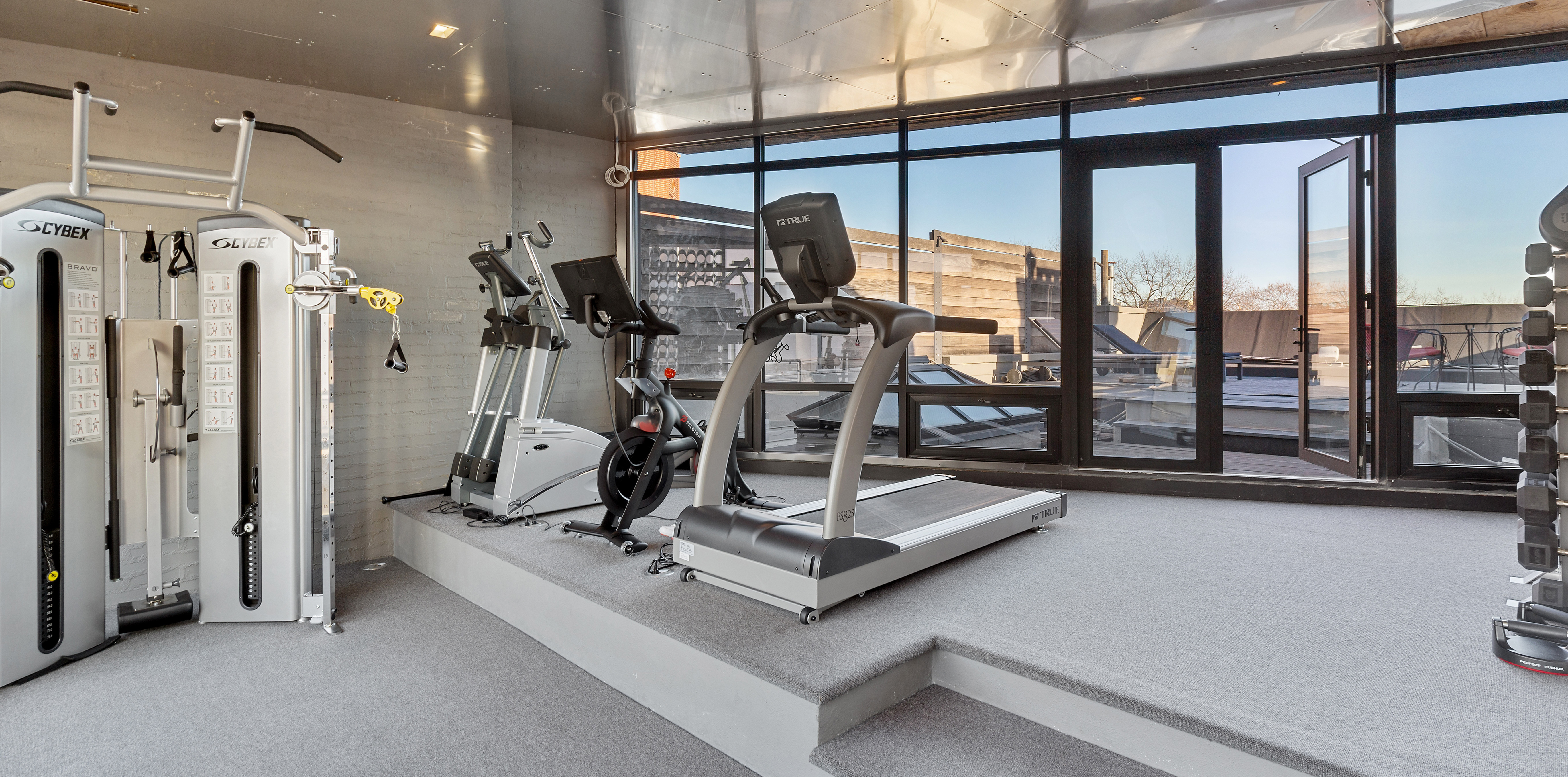 A home gym with a weight machine, stationary bikes, a treadmill, and large glass doors that open to a deck.