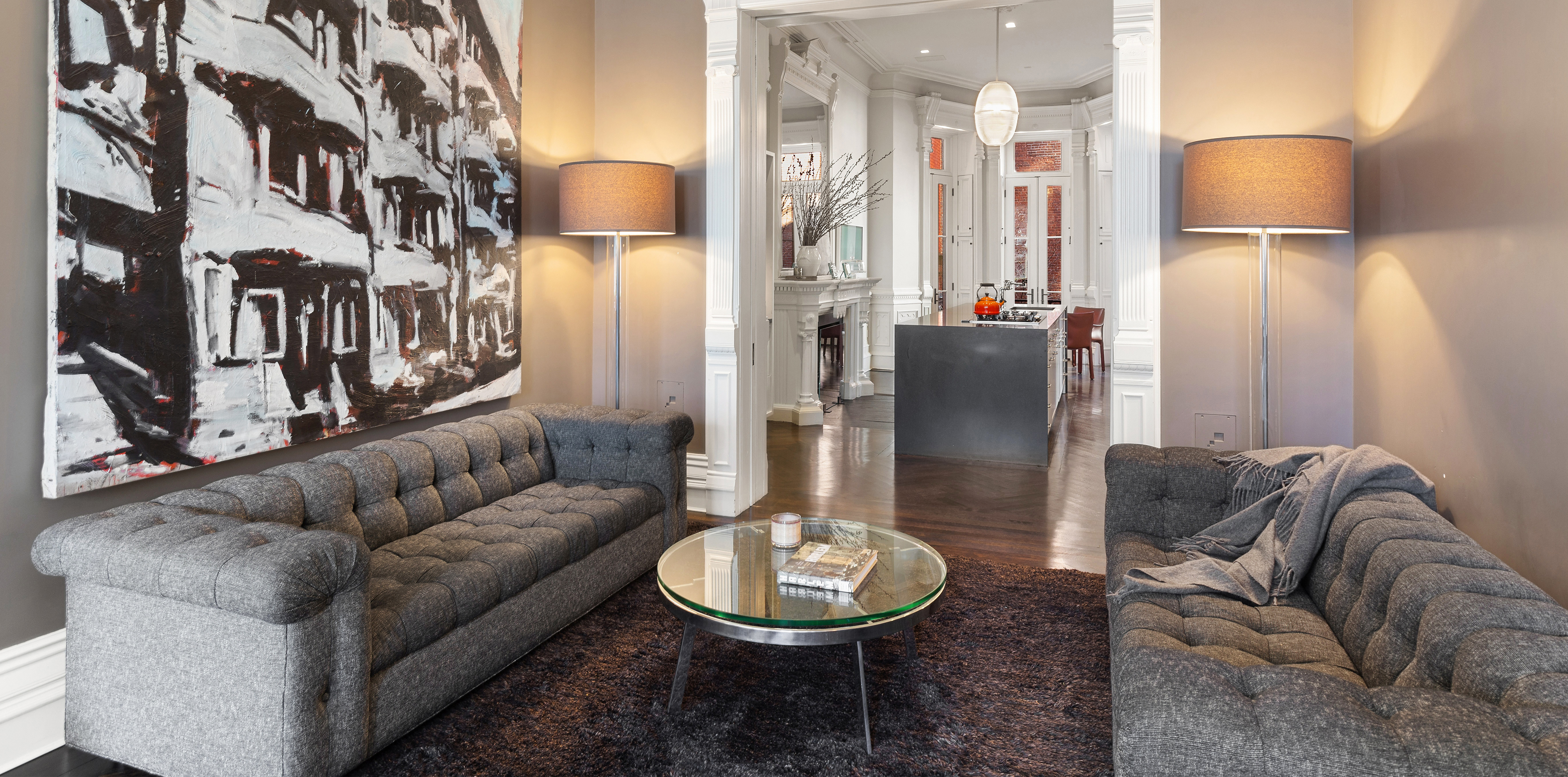 A modern living room with two gray tufted sofas, a round glass coffee table, and a dining area in the background with a white table and chairs on dark hardwood floors.