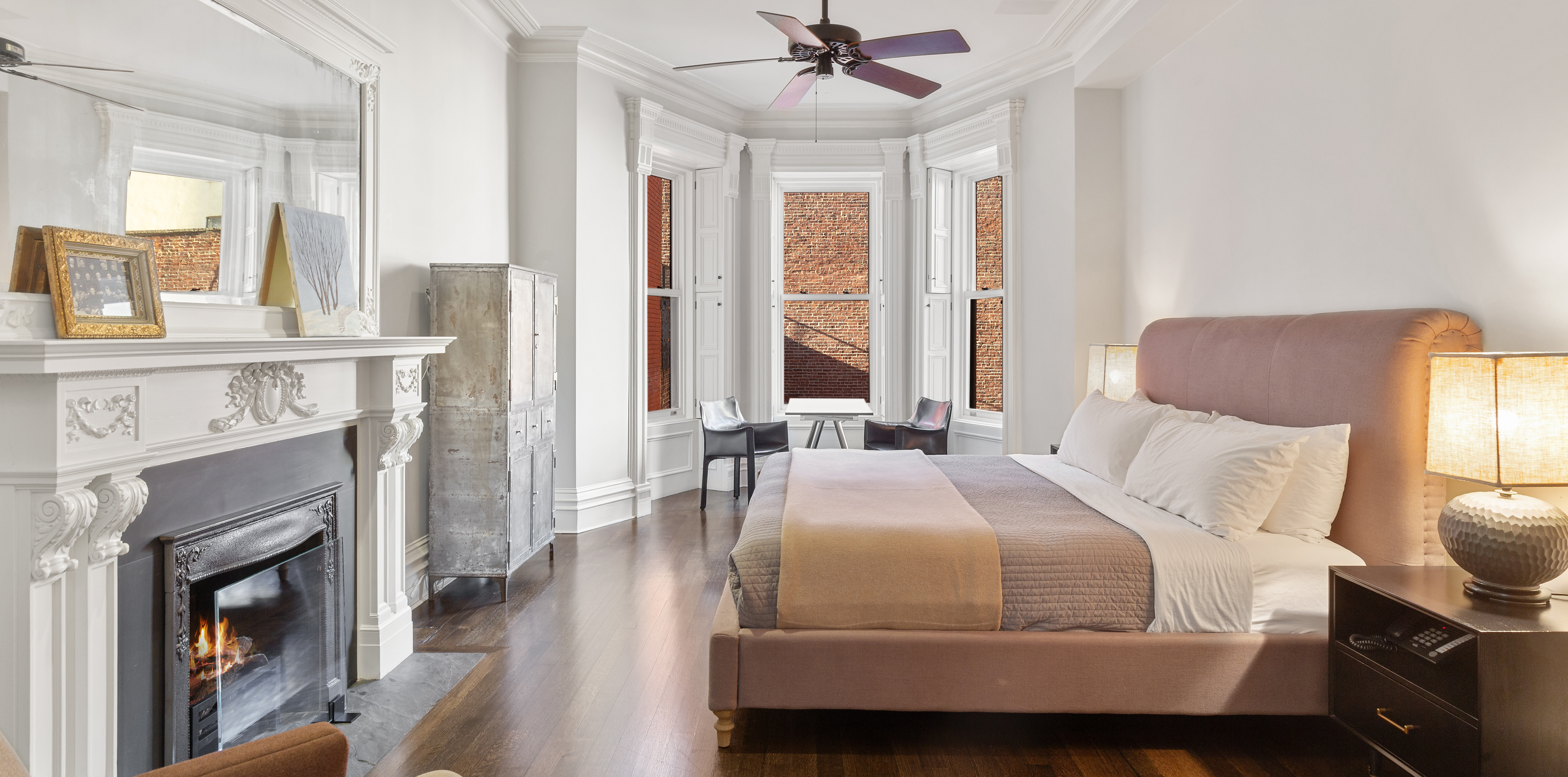 A bedroom with dark wood floors, a lit fireplace, and two chairs sat around a small table by a bay window.