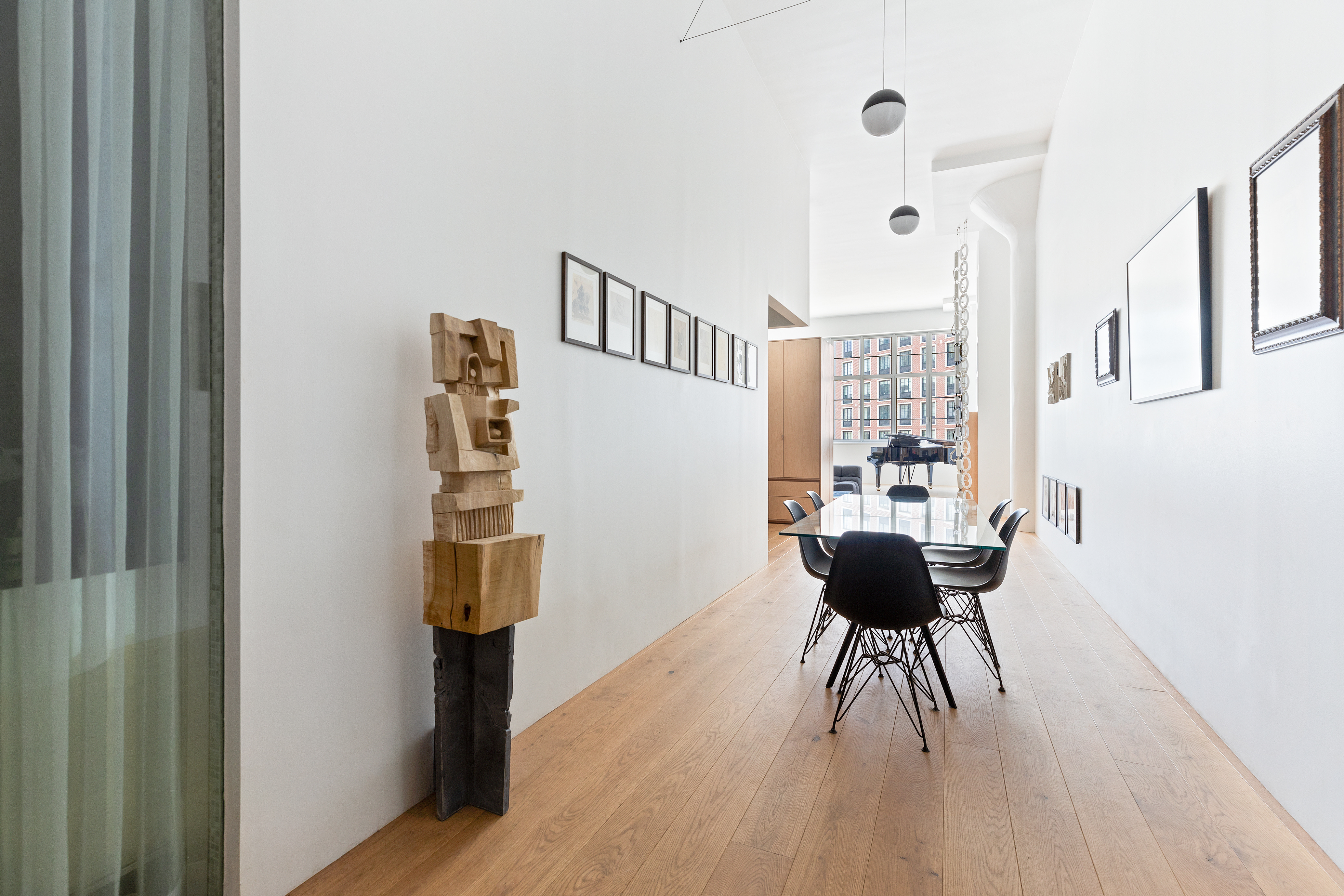 A modern dining room with a long wooden table surrounded by black chairs, a wooden sculpture on the left, framed pictures on white walls, and large windows with sheer curtains letting in natural light.