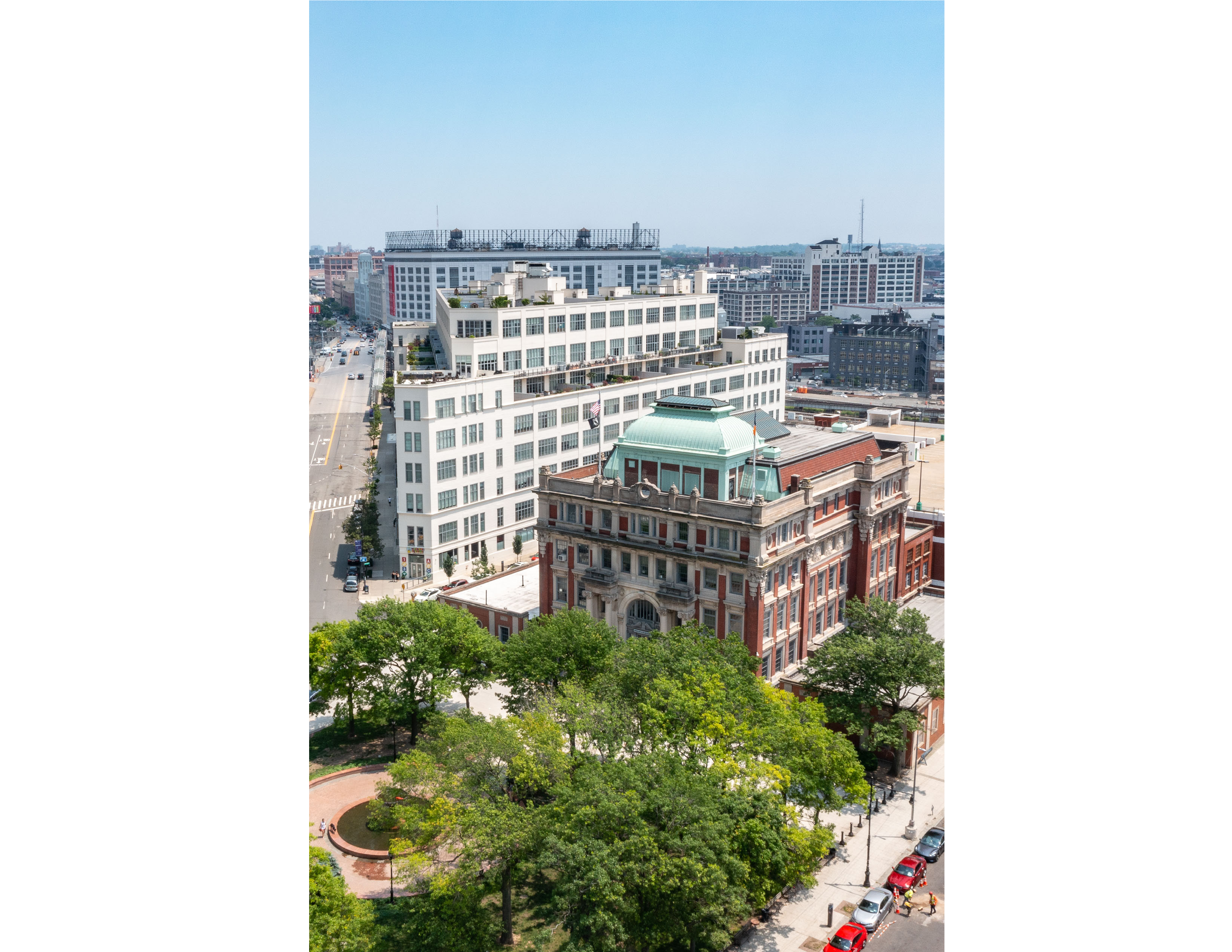 Aerial view of a cityscape with a historic red-brick building topped by a green dome in the foreground, surrounded by trees and greenery, and modern white buildings and roads in the background.