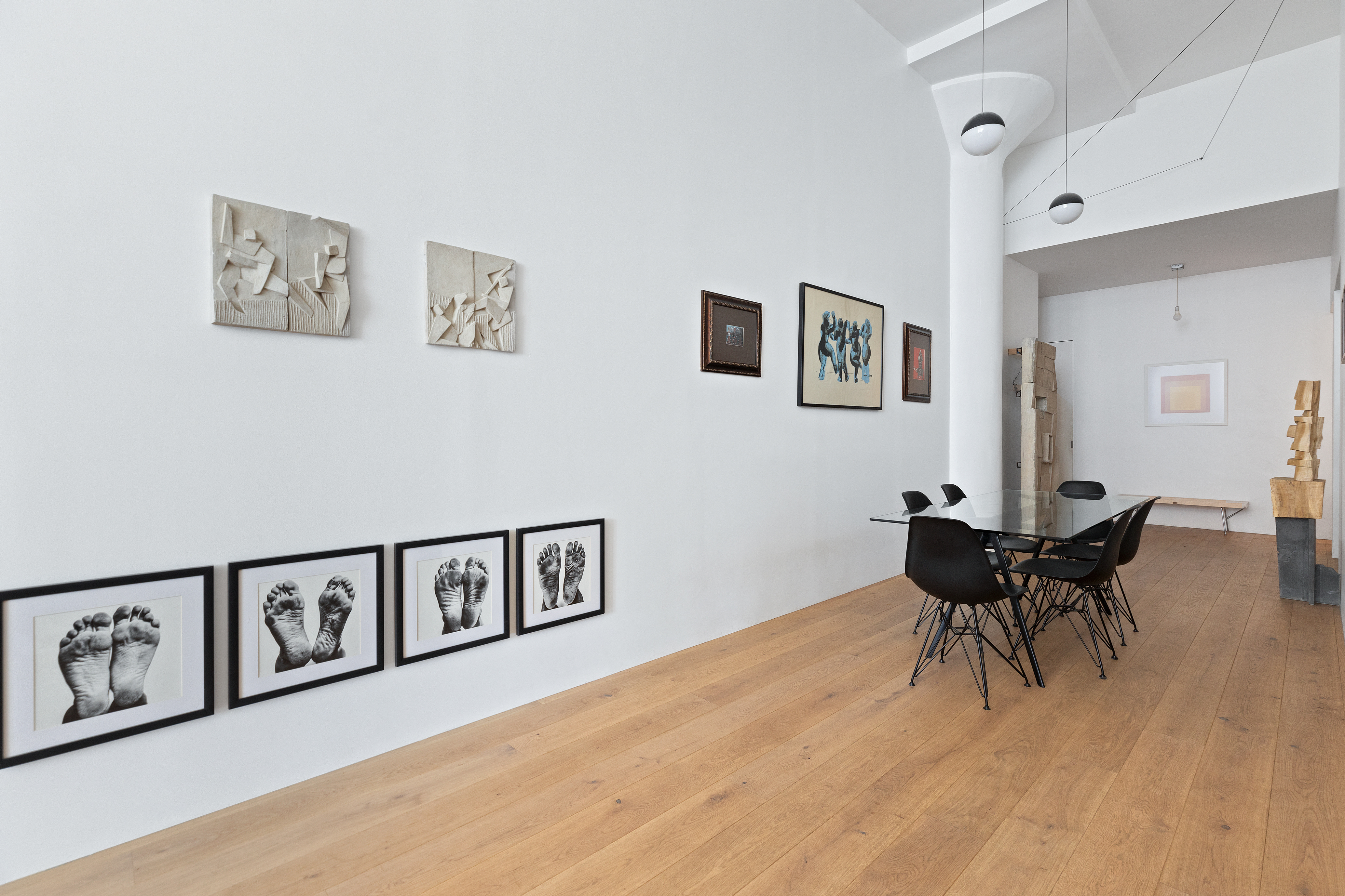 A modern living space with white walls and wooden floors, displaying framed photographs of feet, abstract sculptures, and paintings, with a long black table and black chairs in the center.