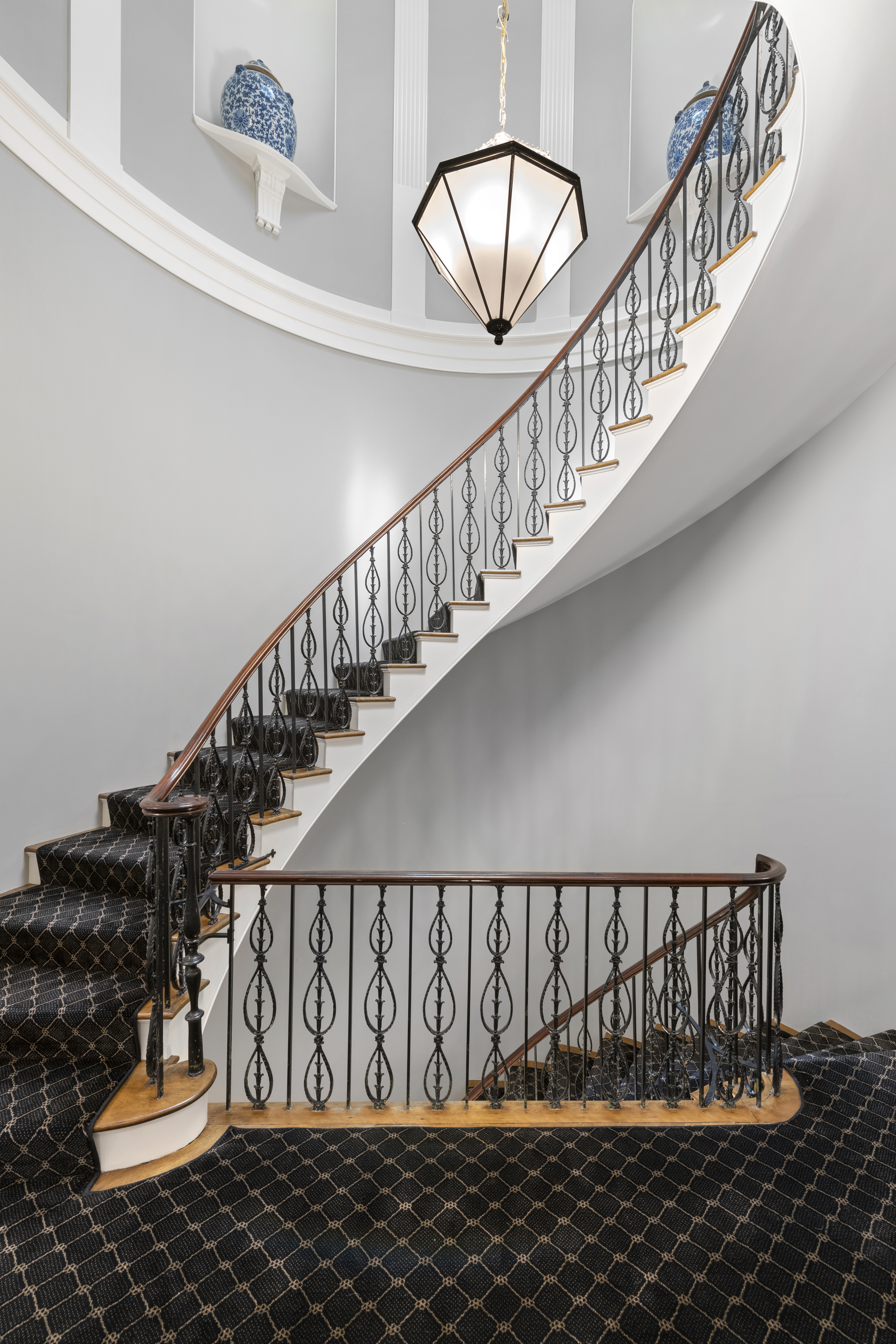 Elegant curved staircase with black patterned carpet and ornate black iron railings, leading to a landing with two blue vases on white display shelves and a geometric pendant light overhead.