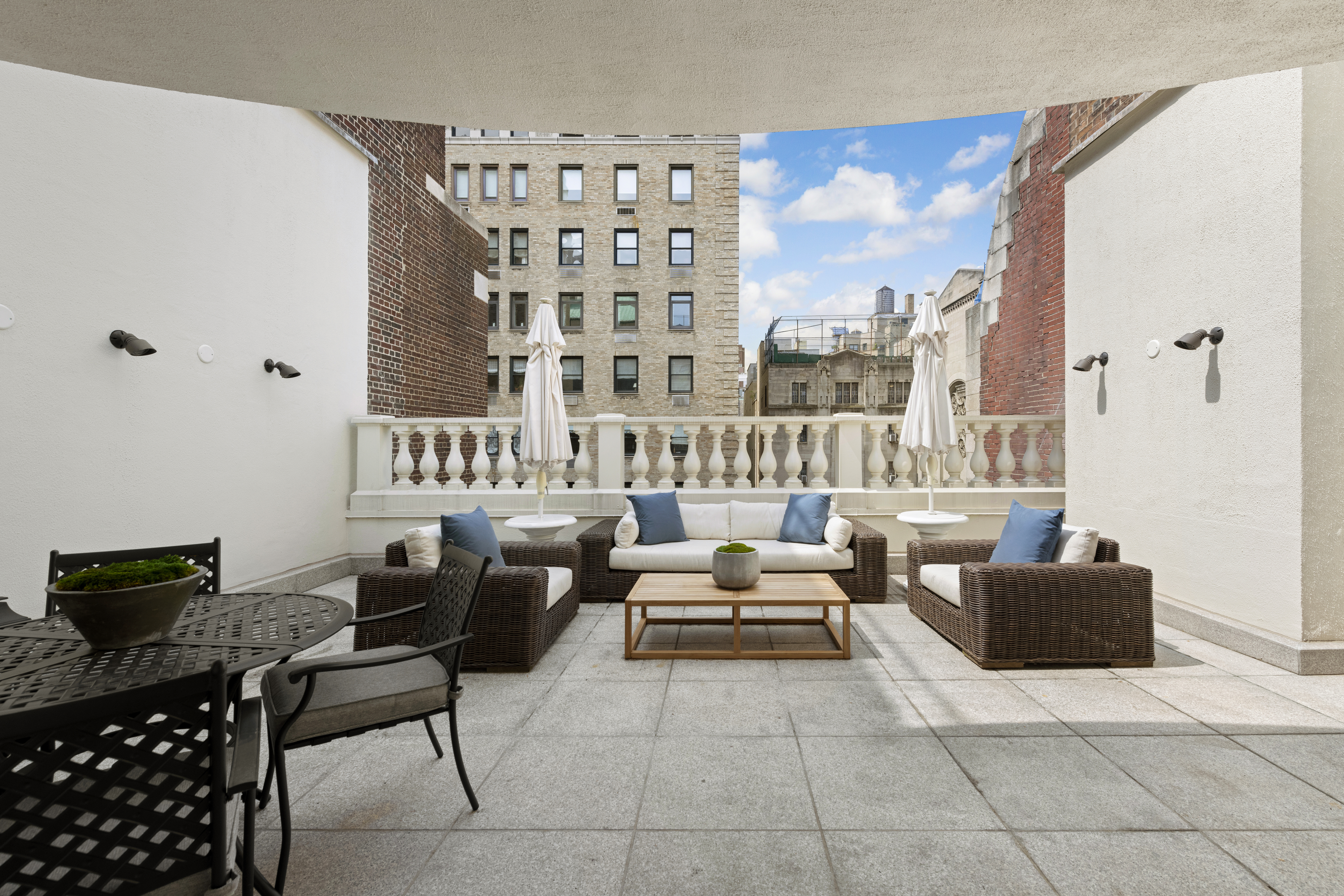 Spacious outdoor patio with wicker furniture, including a sofa and two armchairs with blue and white cushions around a wooden coffee table, flanked by two closed umbrellas and surrounded by buildings.
