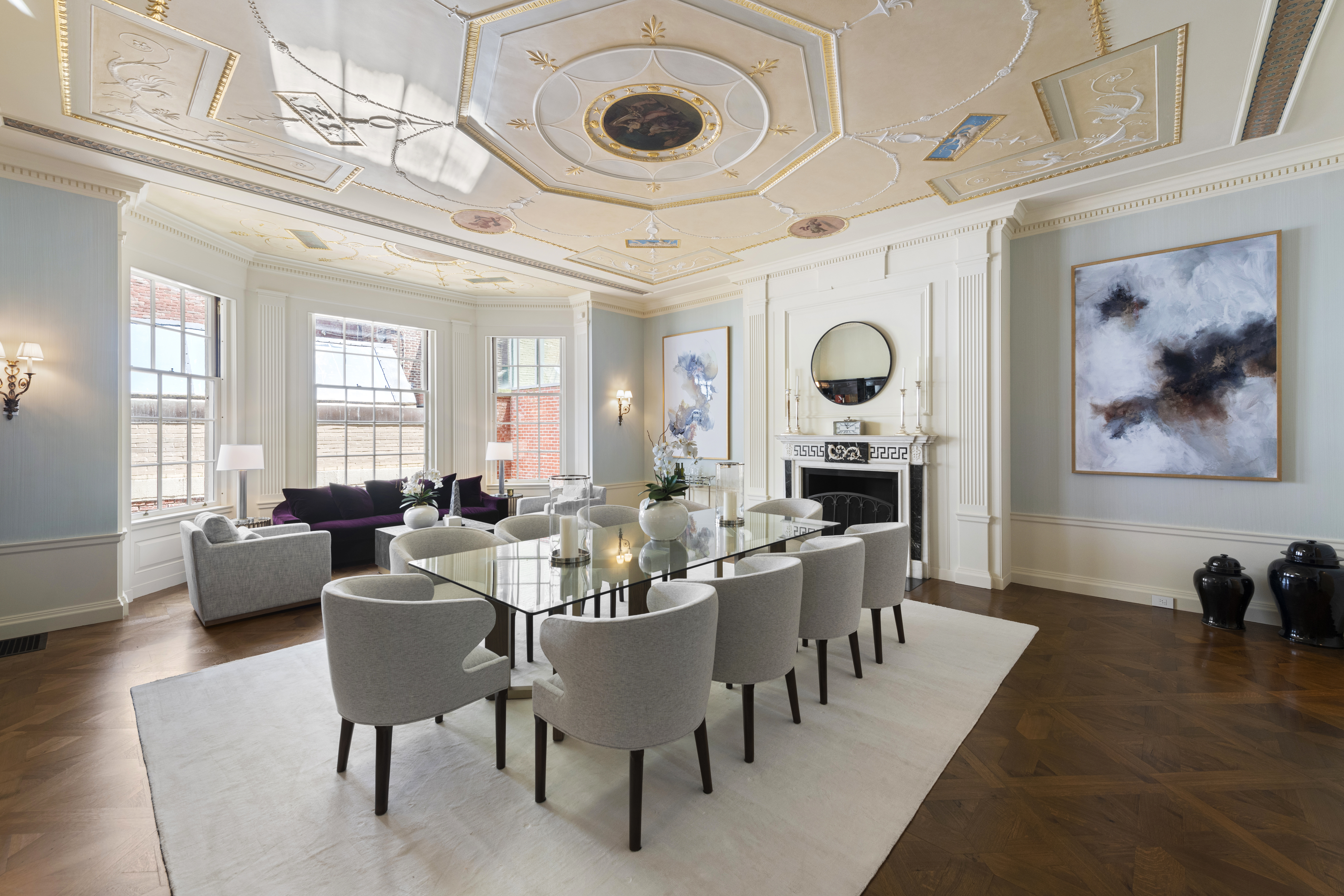 Luxurious dining room with a large glass table and twelve upholstered chairs, ornate ceiling with intricate designs and a central medallion, fireplace with a round mirror above, abstract wall art, and large windows letting in natural light.