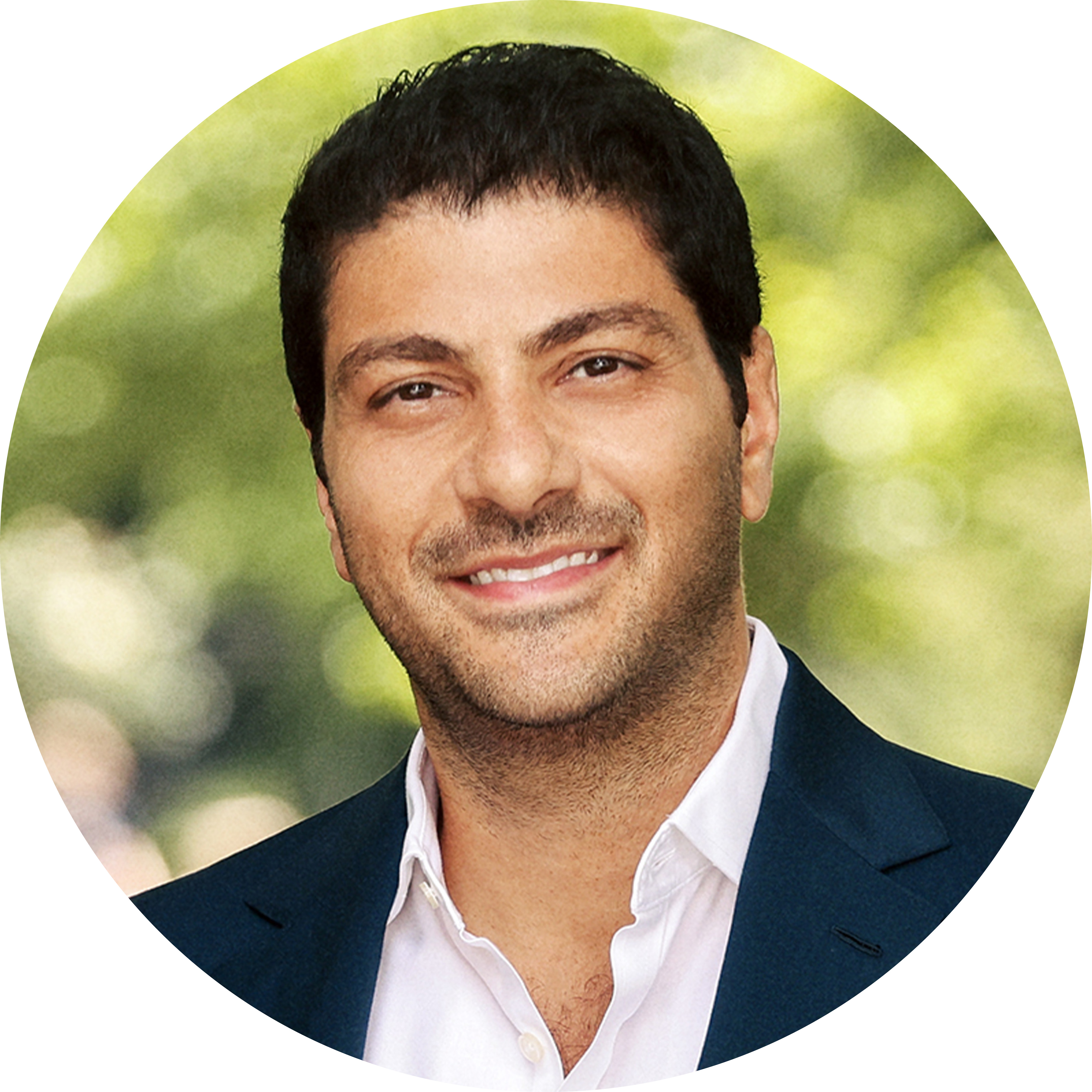 Person wearing a dark suit jacket and white shirt standing outdoors against a blurred green background.