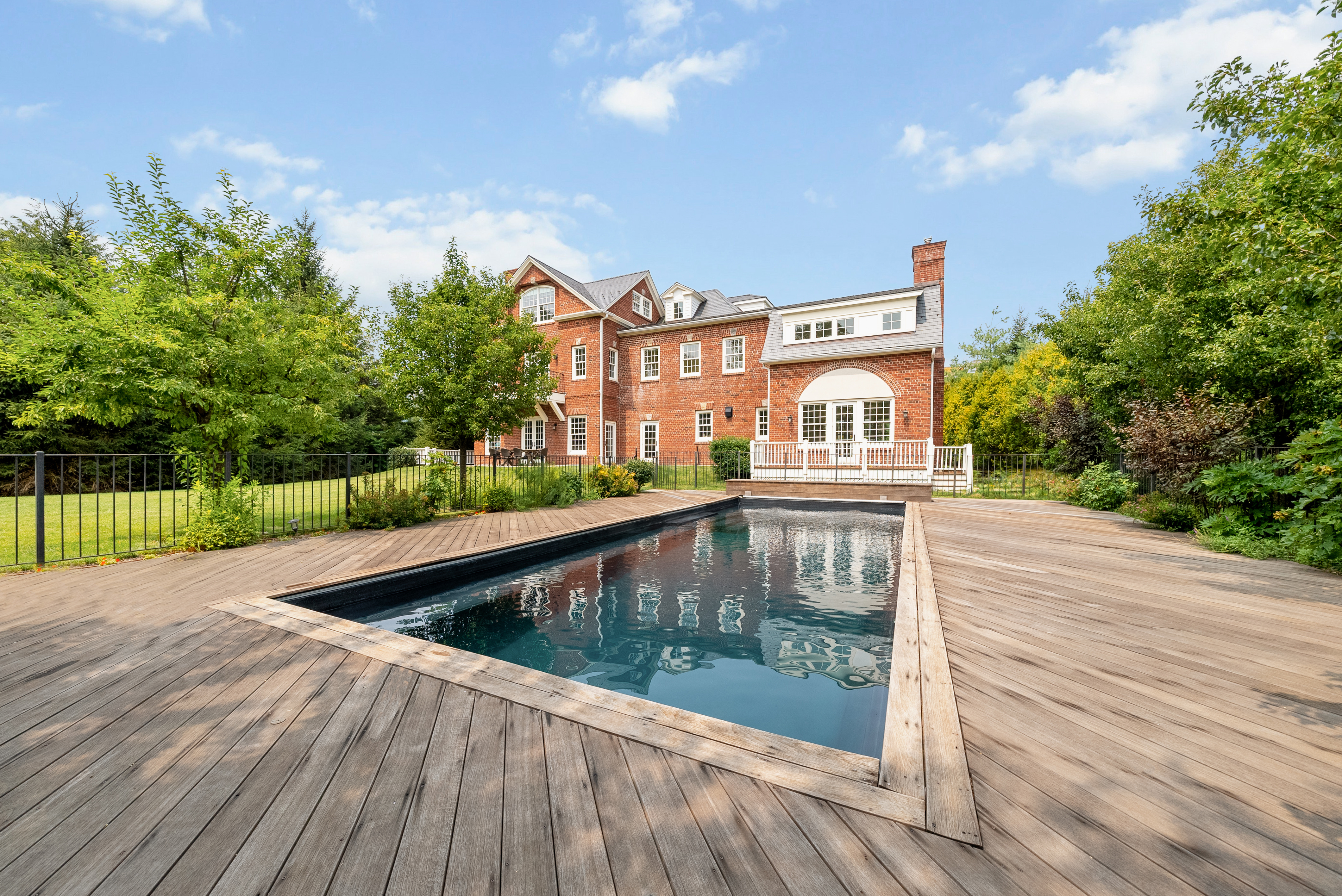 A large rectangular swimming pool with clear water is surrounded by a wooden deck, with a red-brick house and lush green trees in the background.