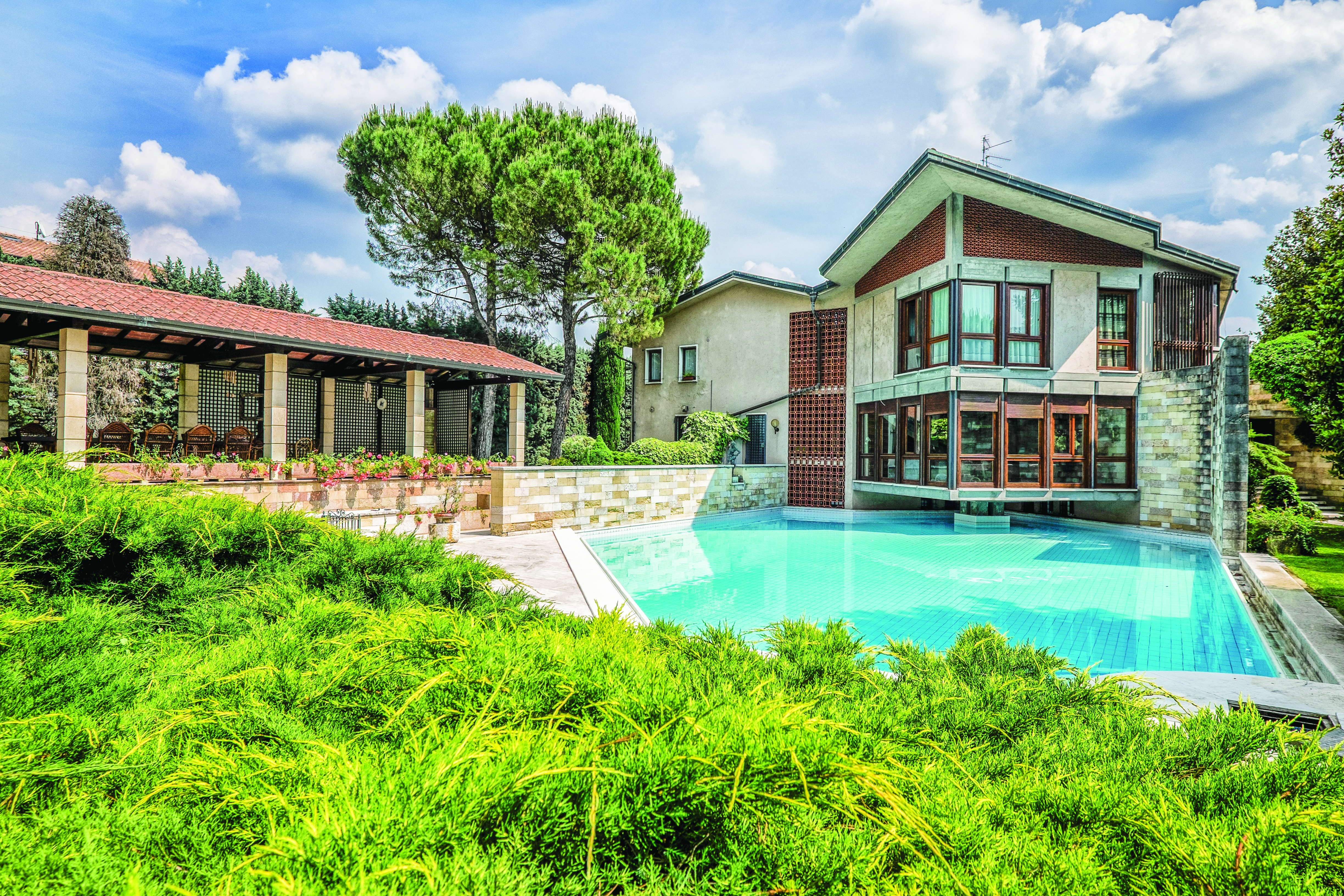 A luxurious modern house with large windows and a spacious patio beside an infinity pool, surrounded by lush greenery and tall trees. The scene is set under a partly cloudy sky.