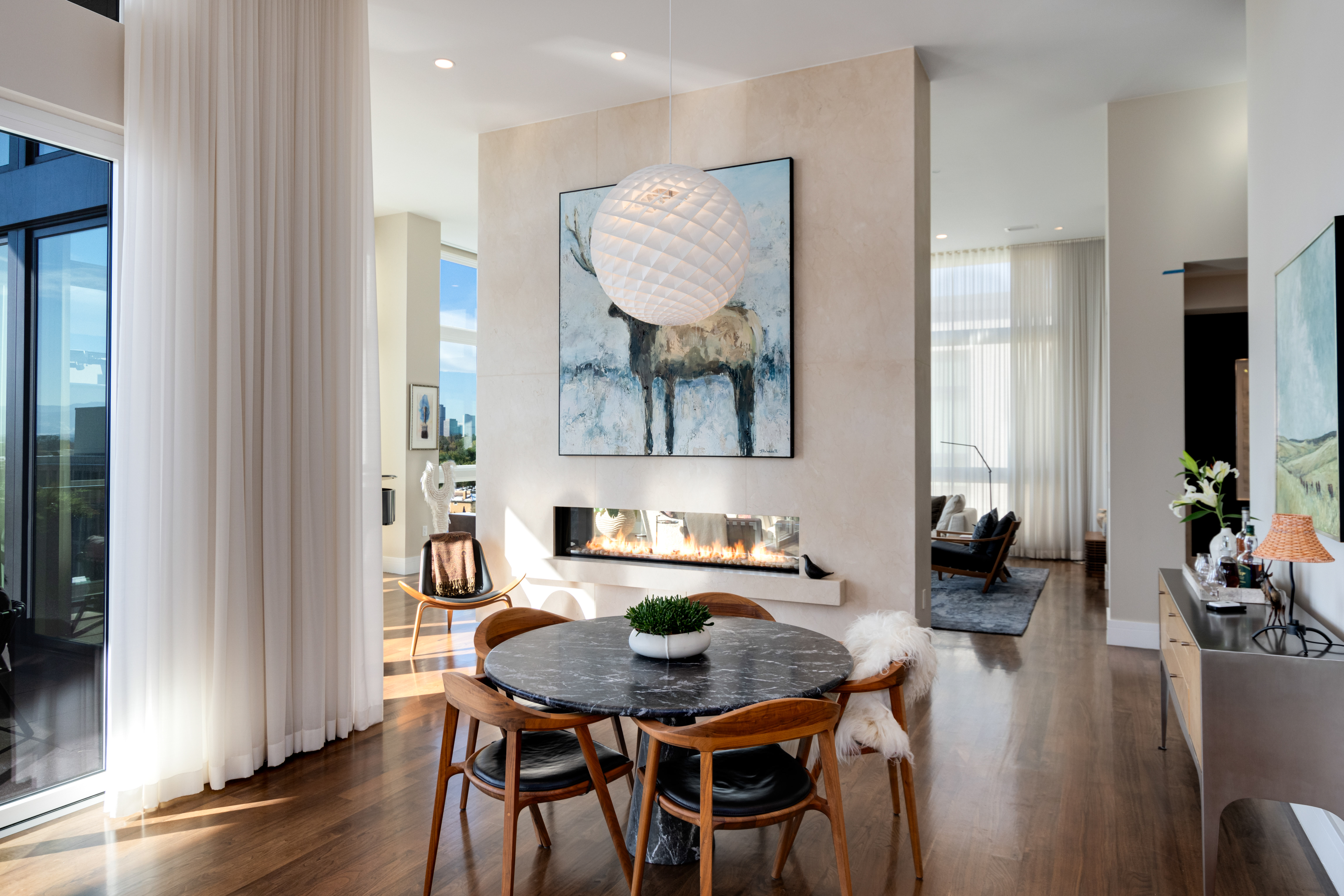 A modern living and dining area with a round black marble table surrounded by wooden chairs, a large painting of a moose above a sleek fireplace, and floor-to-ceiling windows with sheer white curtains allowing natural light to fill the space.