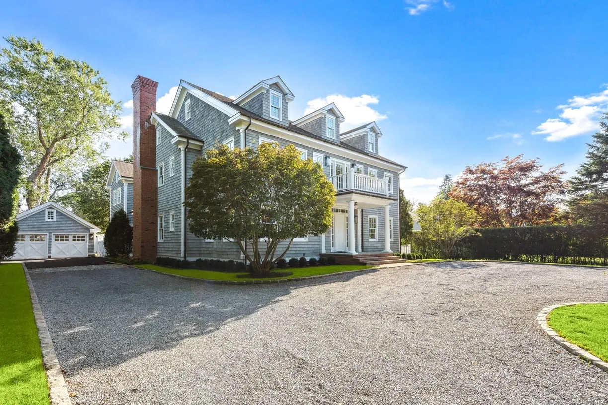 Large two-story gray house with white trim and a prominent brick chimney, set on a well-maintained lawn with a circular driveway and a detached garage in the background, surrounded by trees.