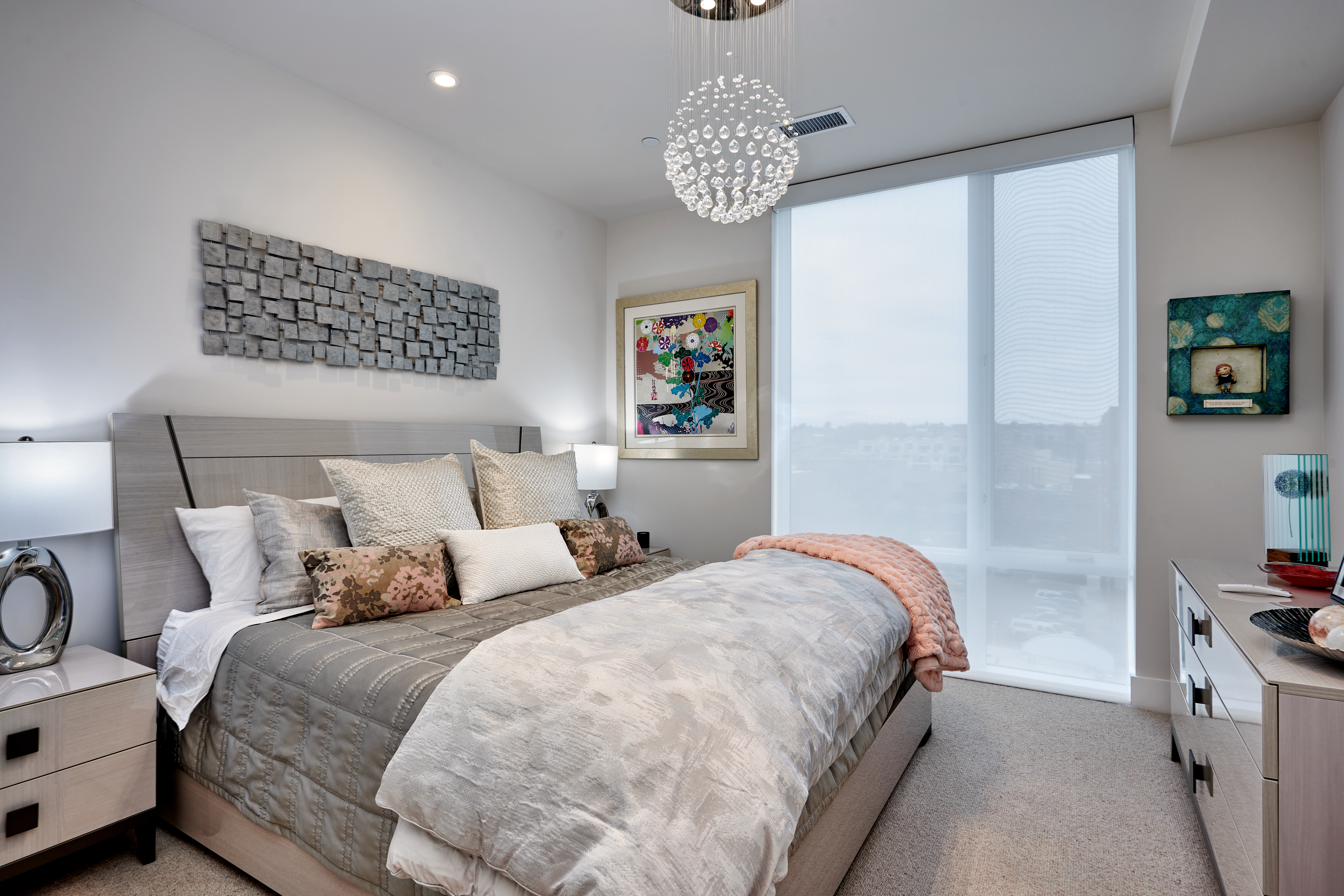 A modern bedroom with a large bed adorned with multiple pillows and blankets, a decorative wall piece above the headboard, a colorful framed artwork, and a spherical chandelier; natural light filters in through a large window with sheer curtains.