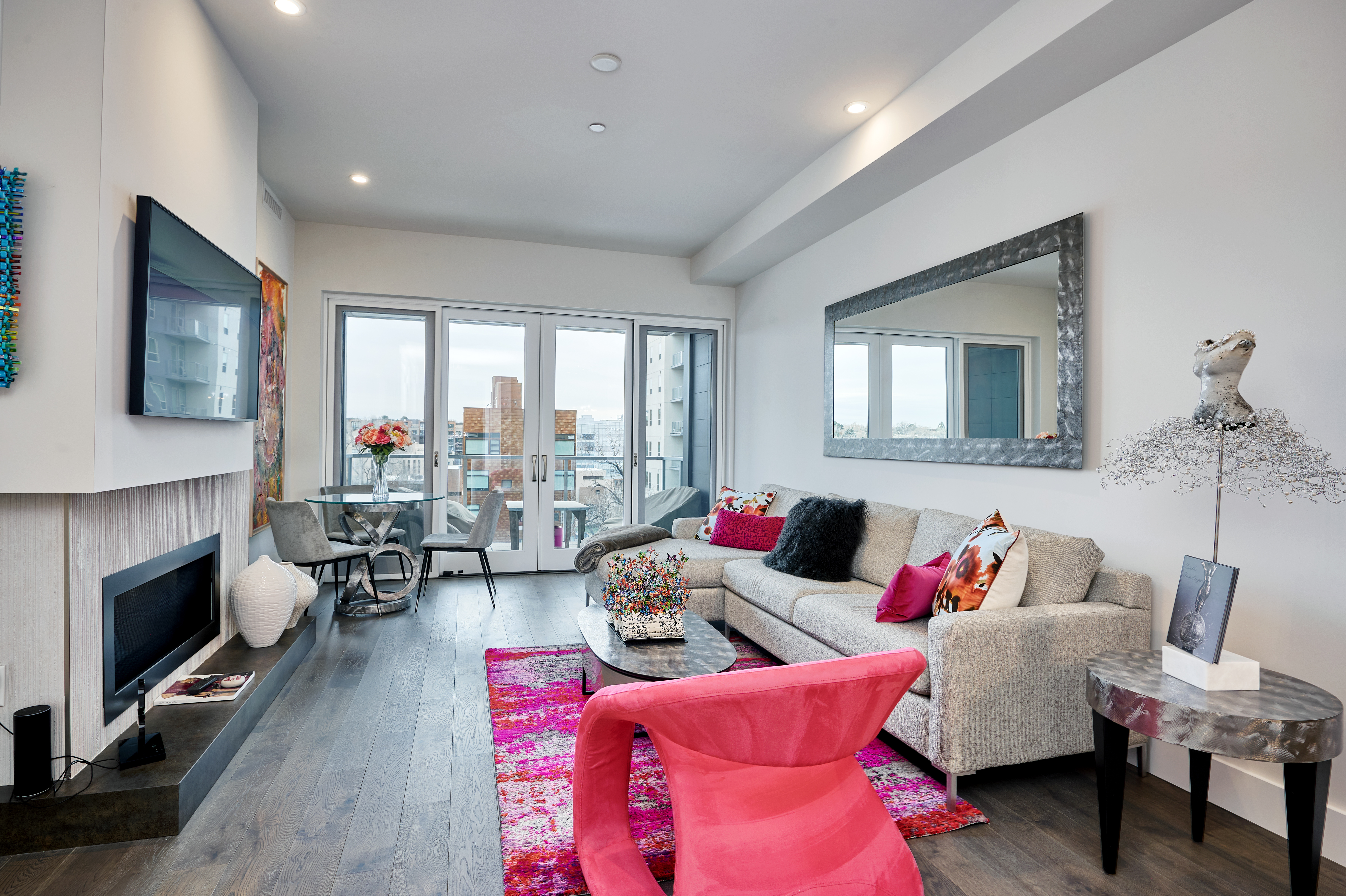 A modern living room with a beige sectional sofa adorned with colorful pillows, a pink chair, and a vibrant pink rug; a large mirror hangs on the right wall, and floor-to-ceiling windows open to a balcony with city views, with a small dining area in the back left corner.
