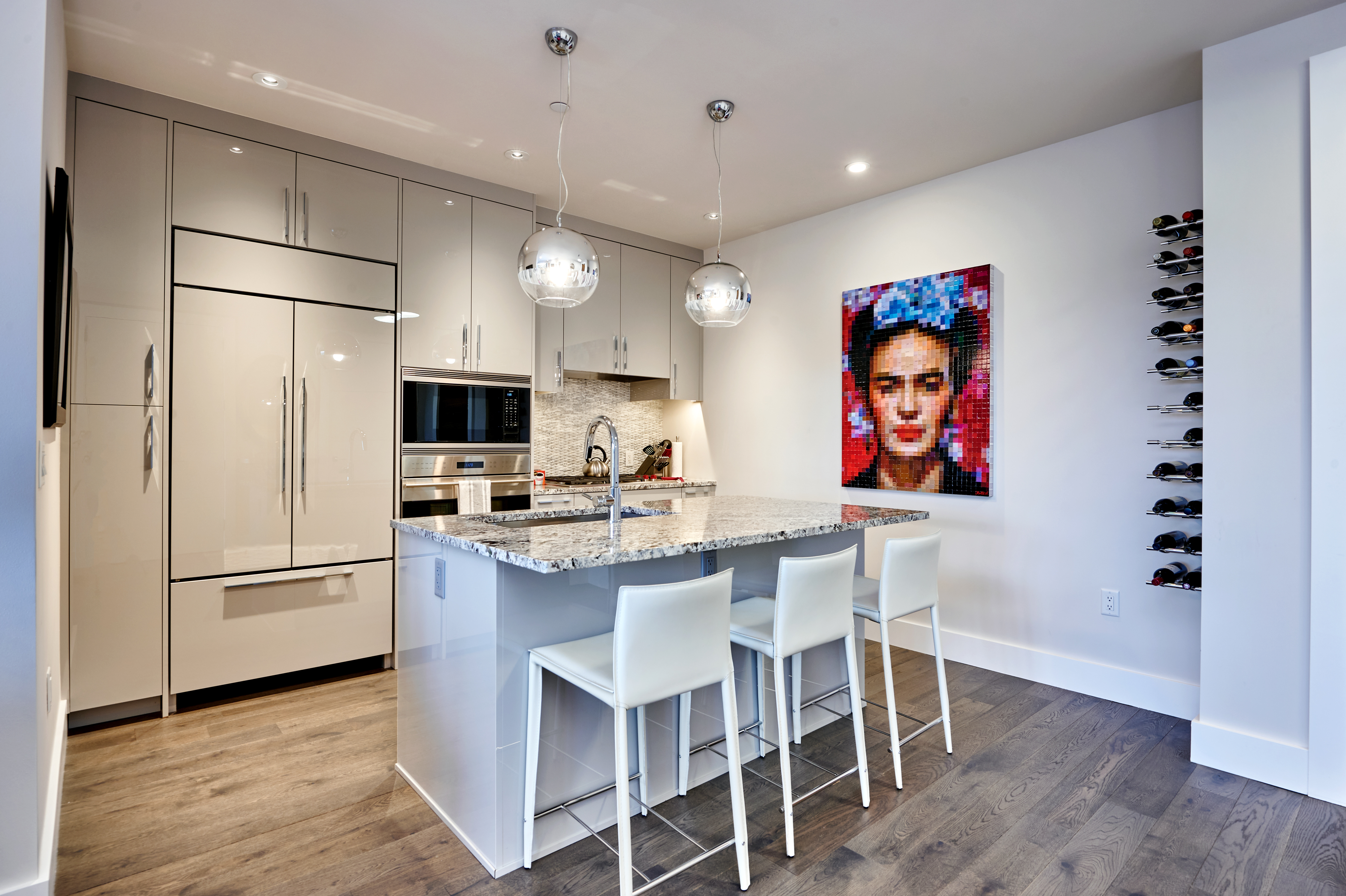 A modern kitchen with a large island featuring a granite countertop, three white bar stools, stainless steel appliances, and two pendant lights; a colorful abstract painting hangs on the wall, and a wine rack filled with bottles is visible to the right.