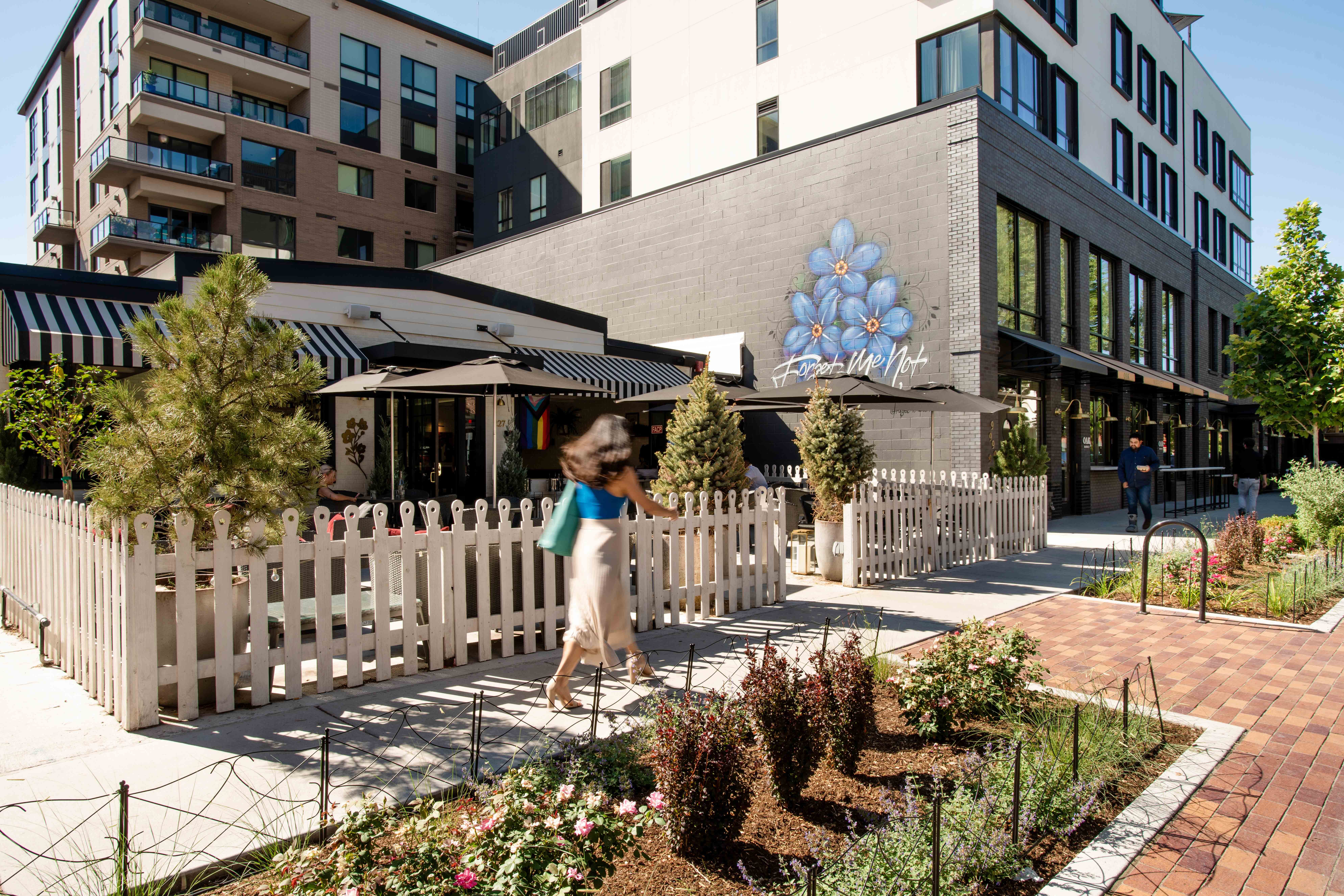 A woman walks past a white picket fence enclosing an outdoor dining area of a restaurant with striped awnings and a blue floral mural; modern apartment buildings and landscaped greenery appear in the background.