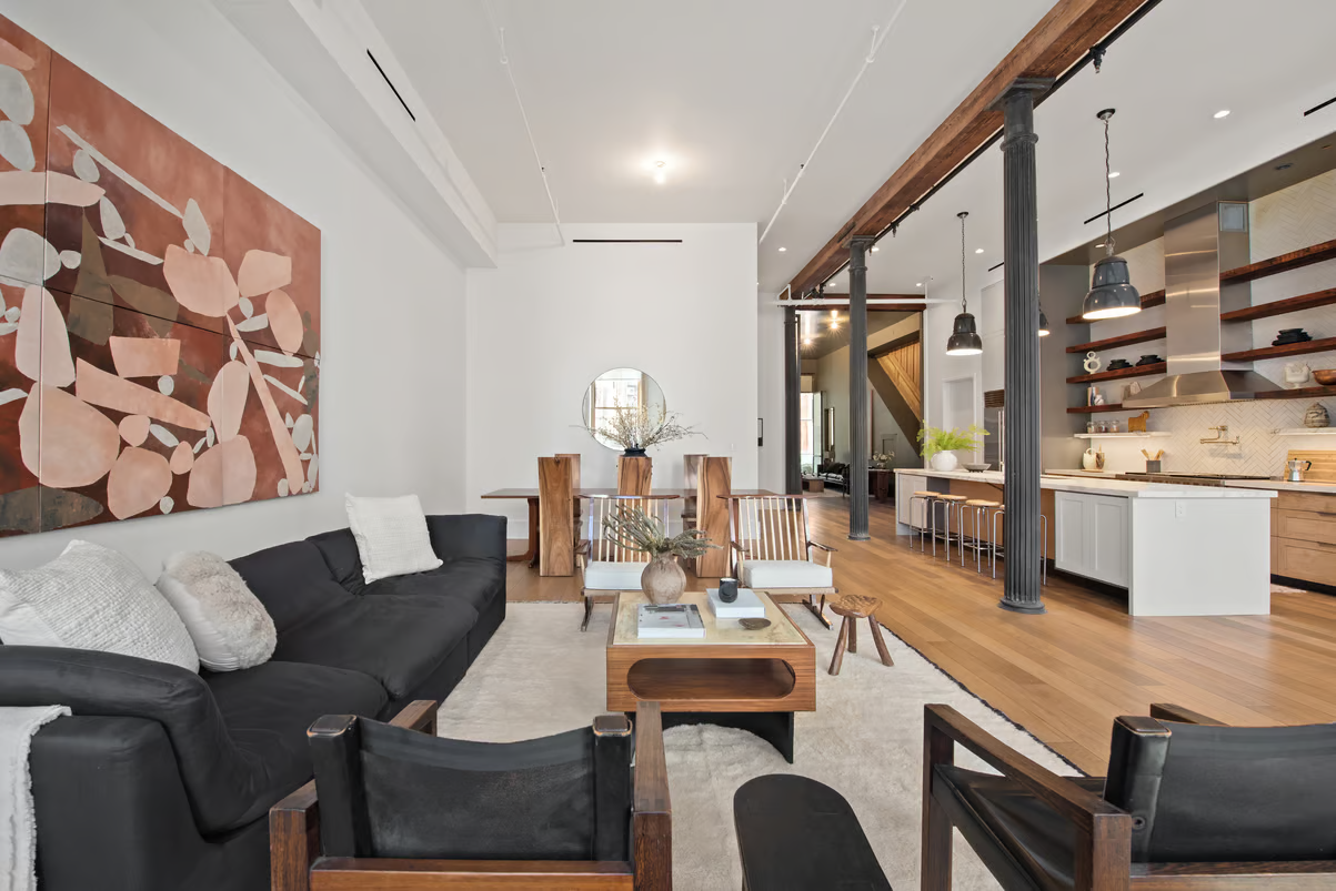 Modern open-concept living room and kitchen with wooden beams, a black sofa, wooden coffee table, abstract wall art, white cabinets, and a large kitchen island with pendant lighting.

