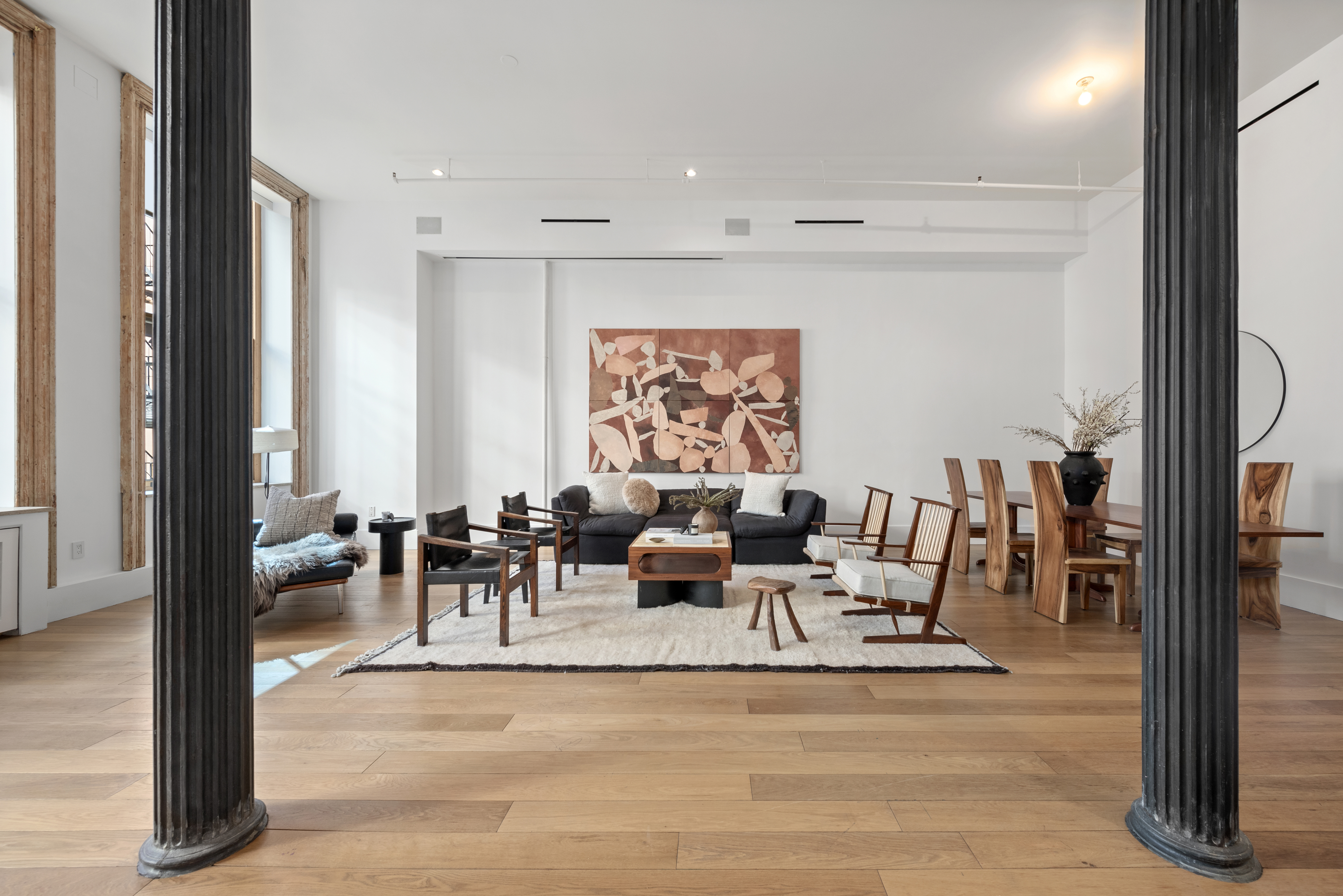 A spacious, modern living room with wooden floors, a black sofa, assorted chairs, a coffee table on a white rug, and a large abstract painting on the wall; the room is framed by two black columns and illuminated by natural light from large windows.