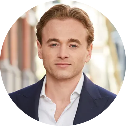 A person with short, light brown hair wearing a white shirt and dark blue blazer stands outdoors, with blurred buildings and trees in the background.