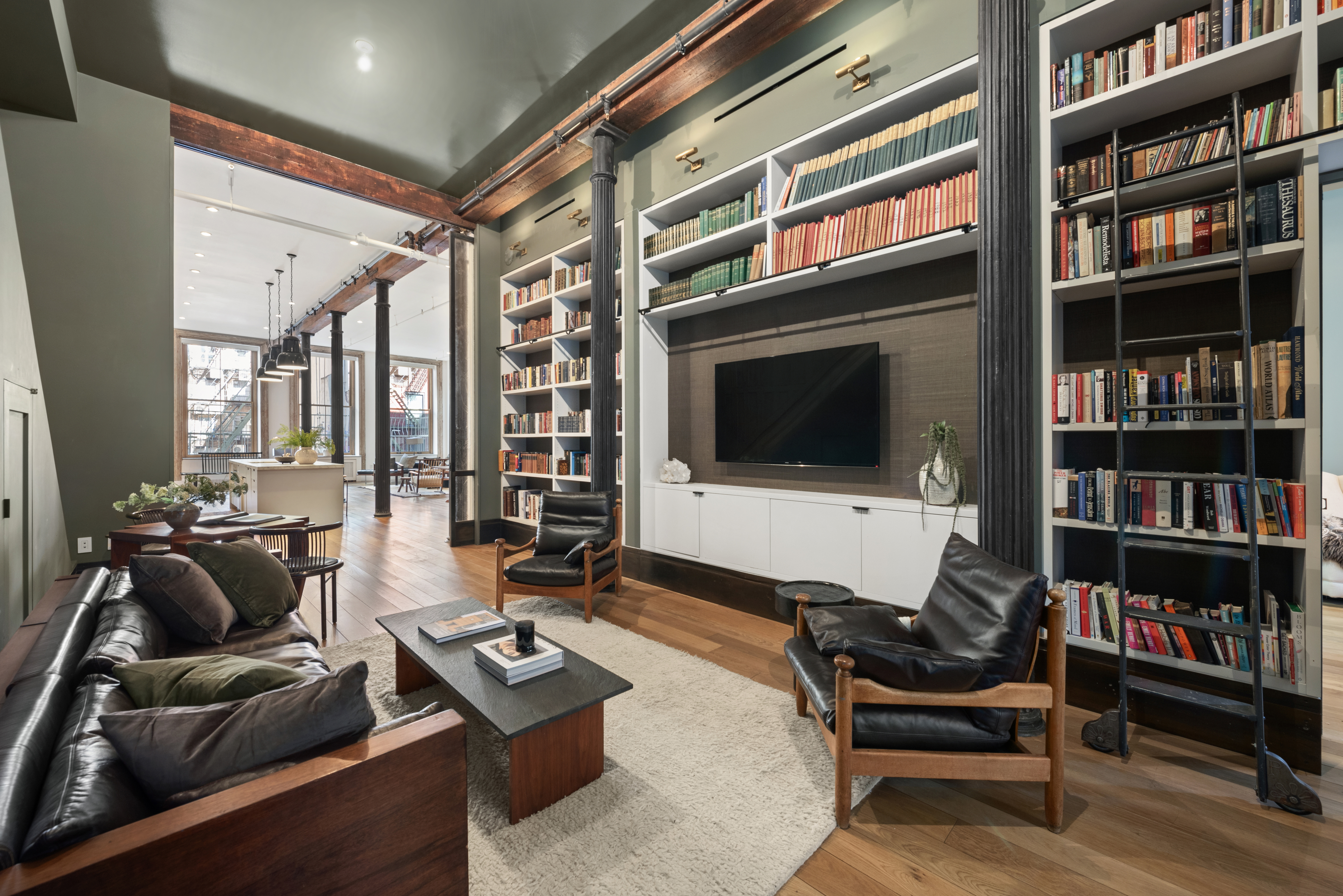 A spacious, modern living room with high ceilings, a wall-mounted flat-screen TV surrounded by built-in bookshelves filled with books, two black leather armchairs, a wooden coffee table, and a dark brown sofa with cushions; an open kitchen is visible in the background.