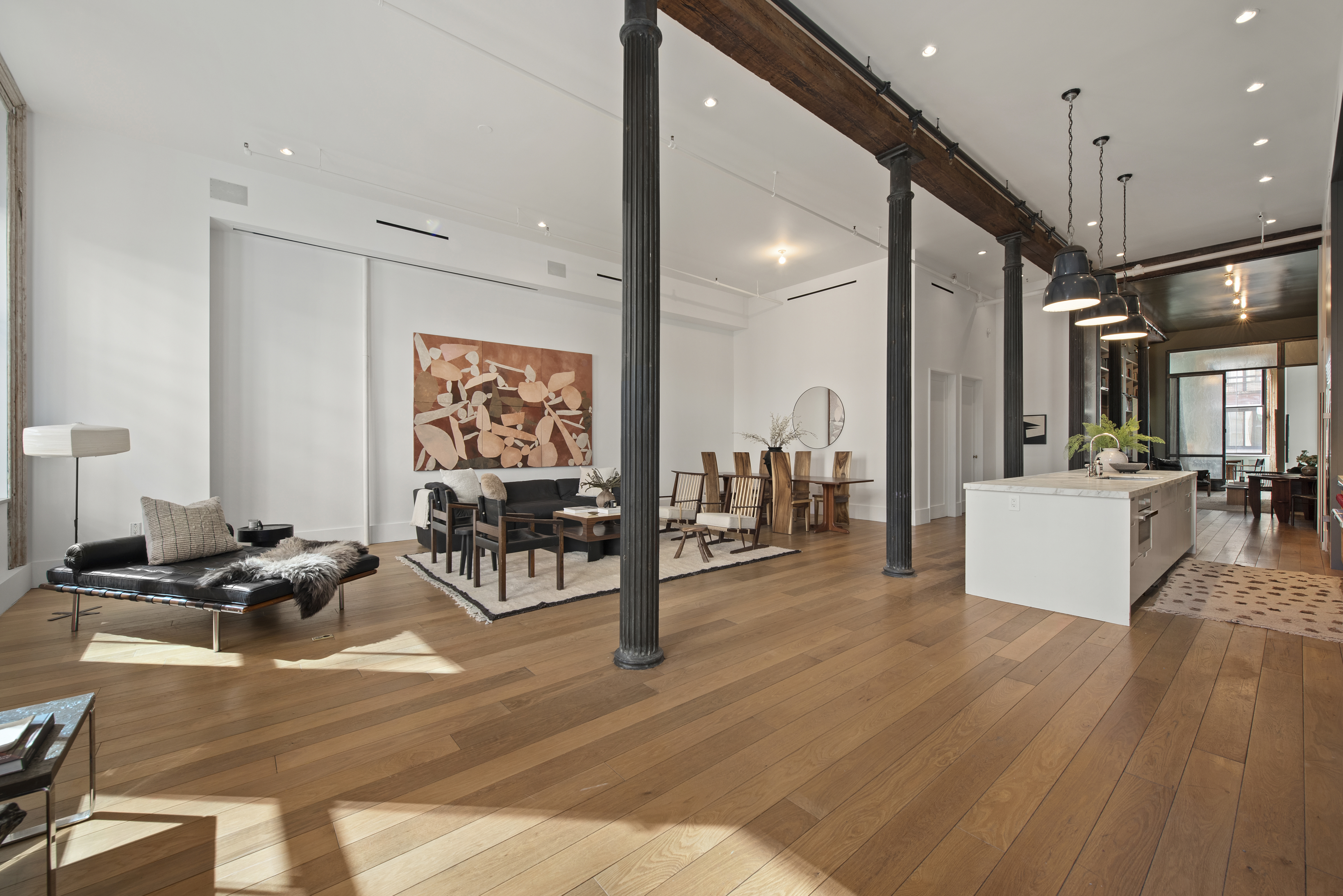 A spacious, modern open-concept living area with wooden floors, black lounge furniture, a large abstract painting, a dining table with chairs, and an island kitchen with pendant lights; natural light streams in through large windows at the far end.