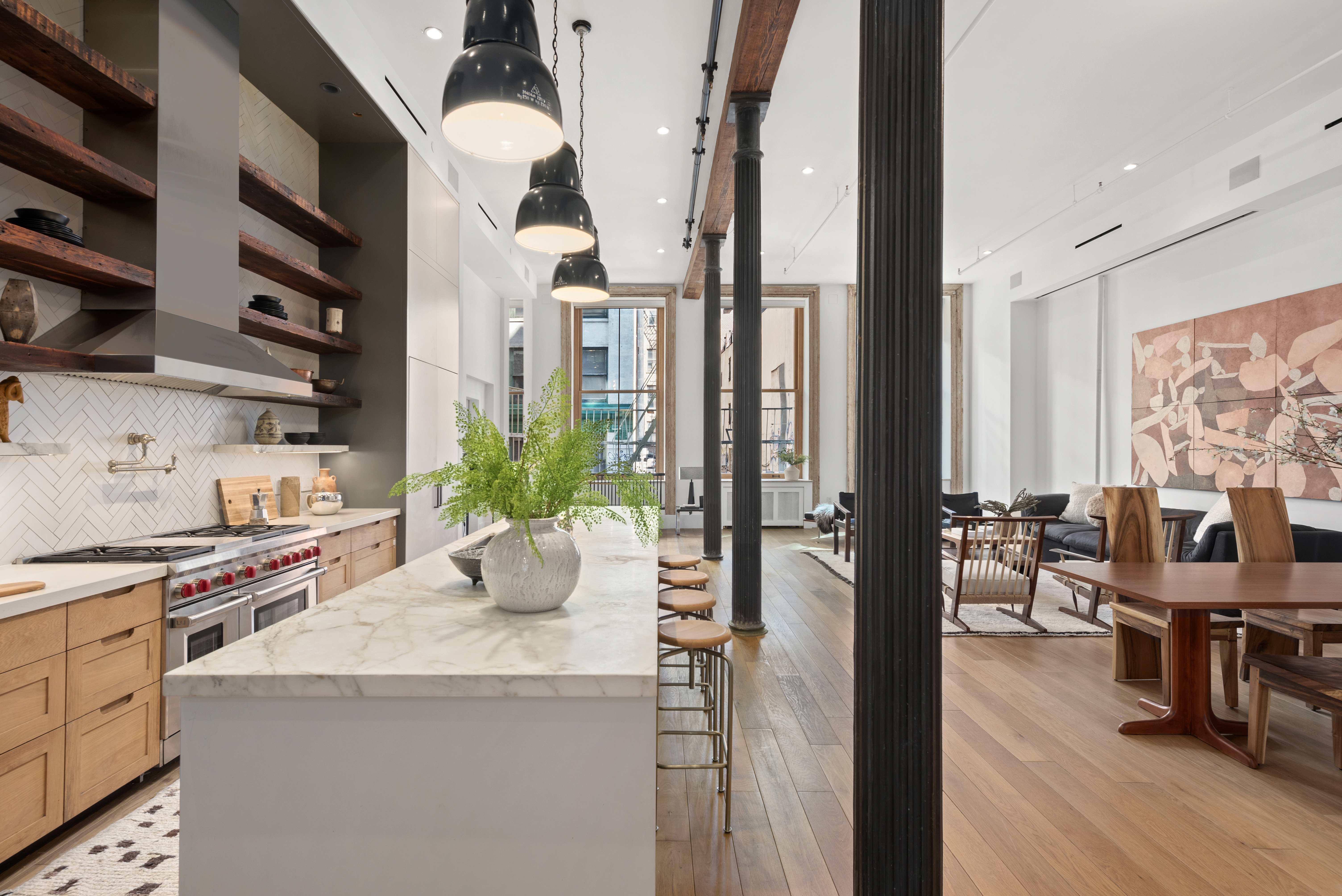 A modern, open-concept kitchen and dining area with wooden floors, a marble island topped with a potted fern, bar stools, black pendant lights, and a spacious living area in the background illuminated by large windows.